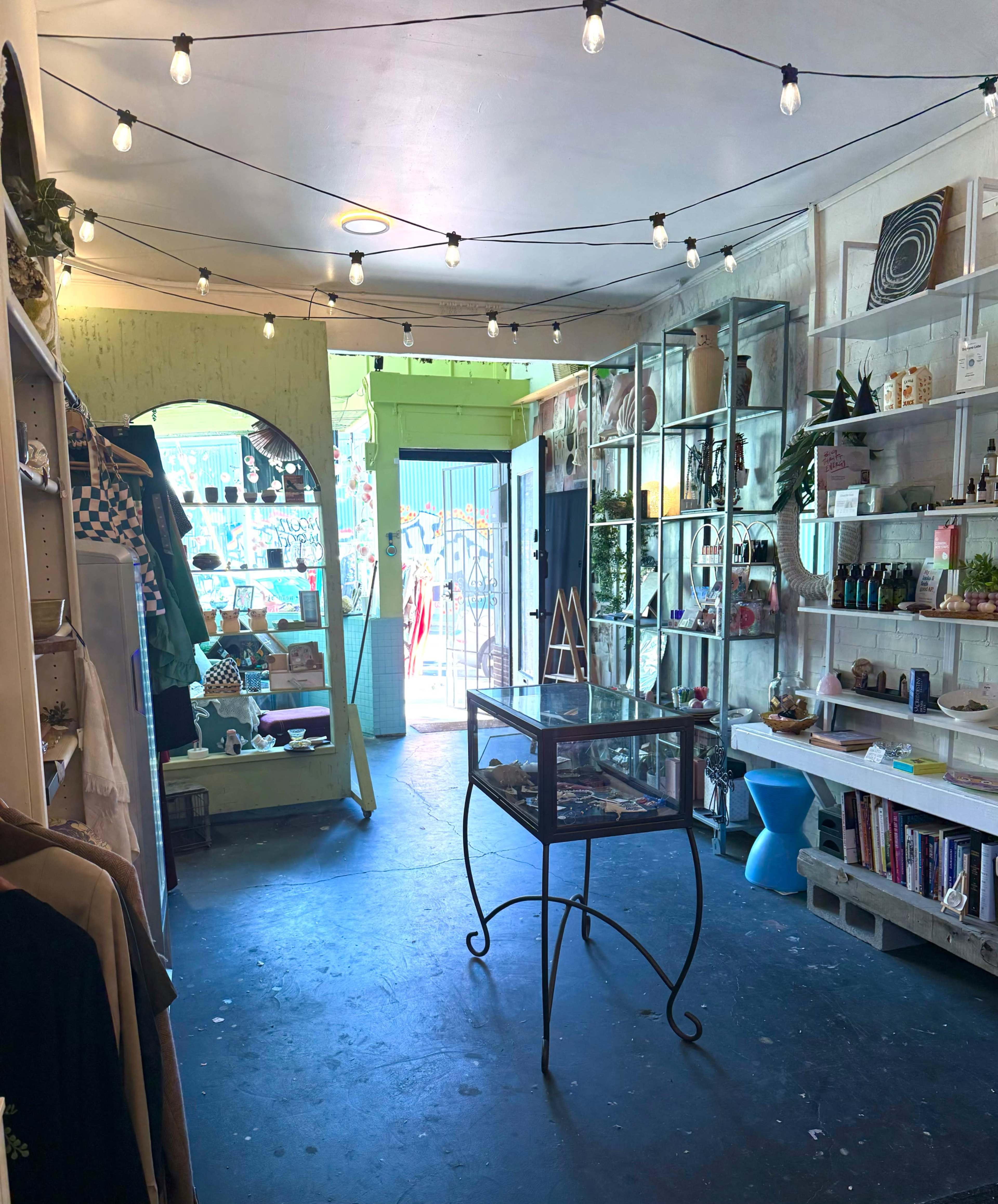 The image shows an interior of a shop with a display case in the center, shelves lined with various items, and fairy lights hanging from the ceiling.