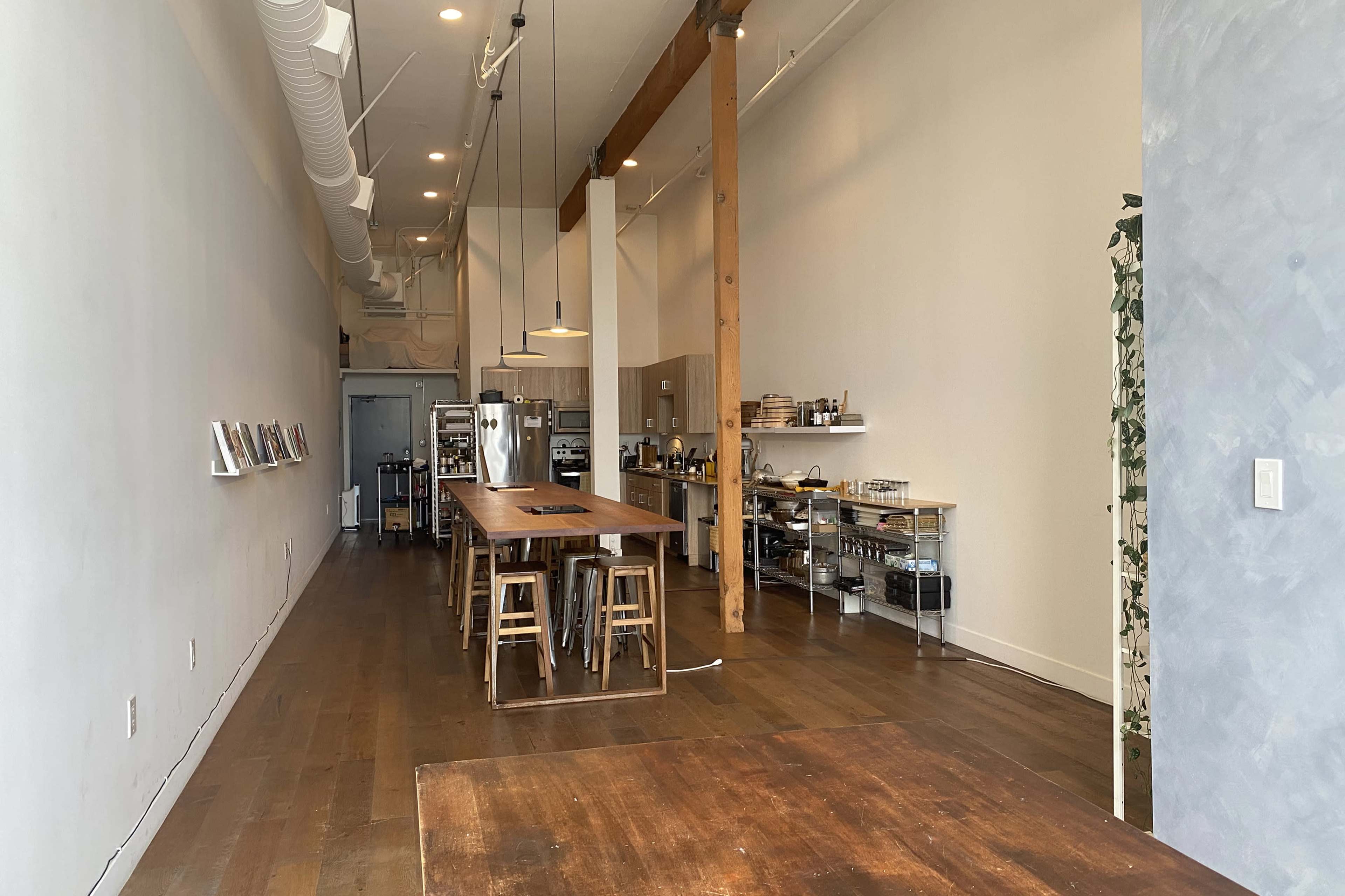 A spacious interior of a modern kitchen with wooden flooring, a long table surrounded by stools, and shelving units along the walls.
