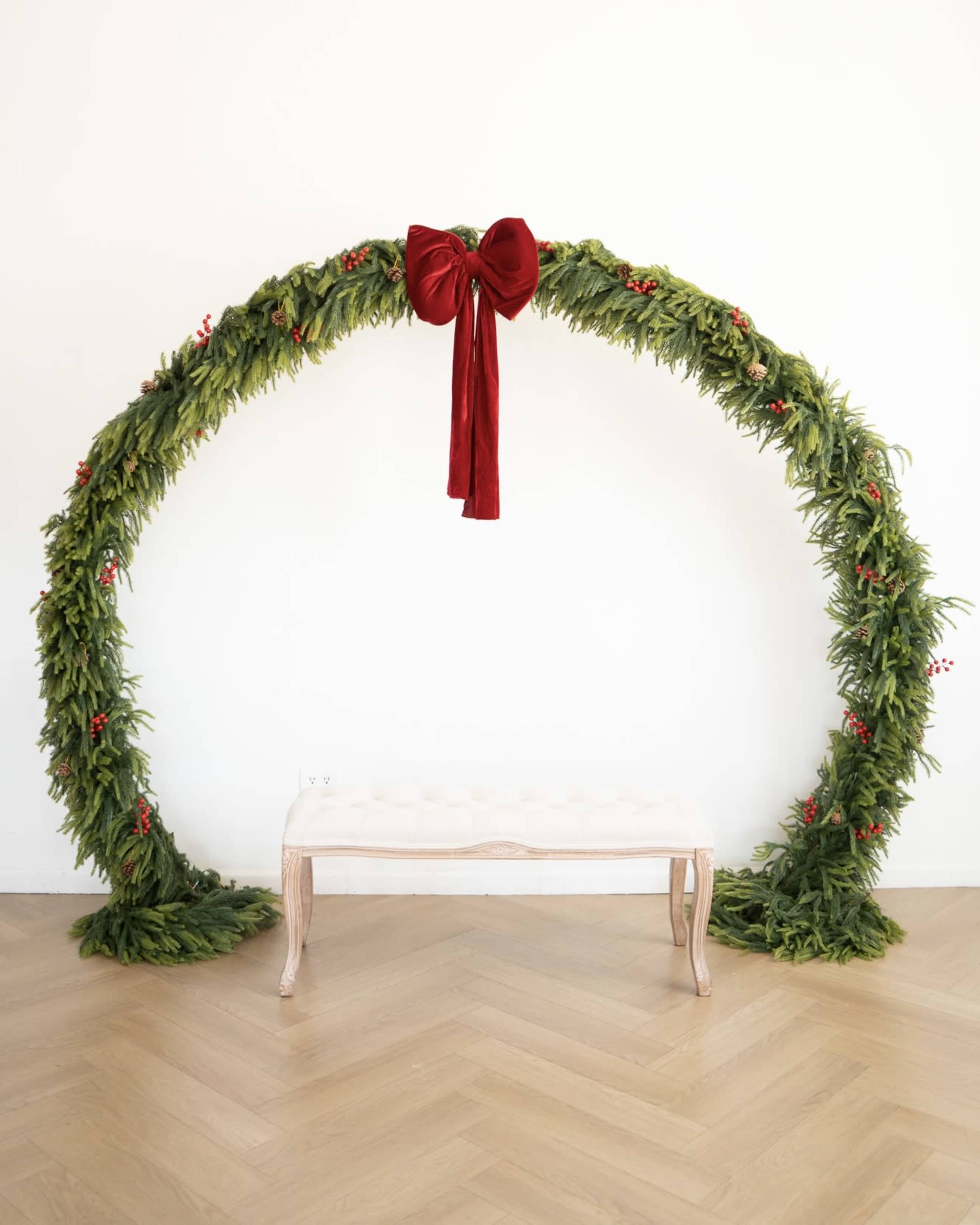 A lush green arch made of foliage and decorated with a large red bow stands above a simple light-colored bench against a plain white wall.