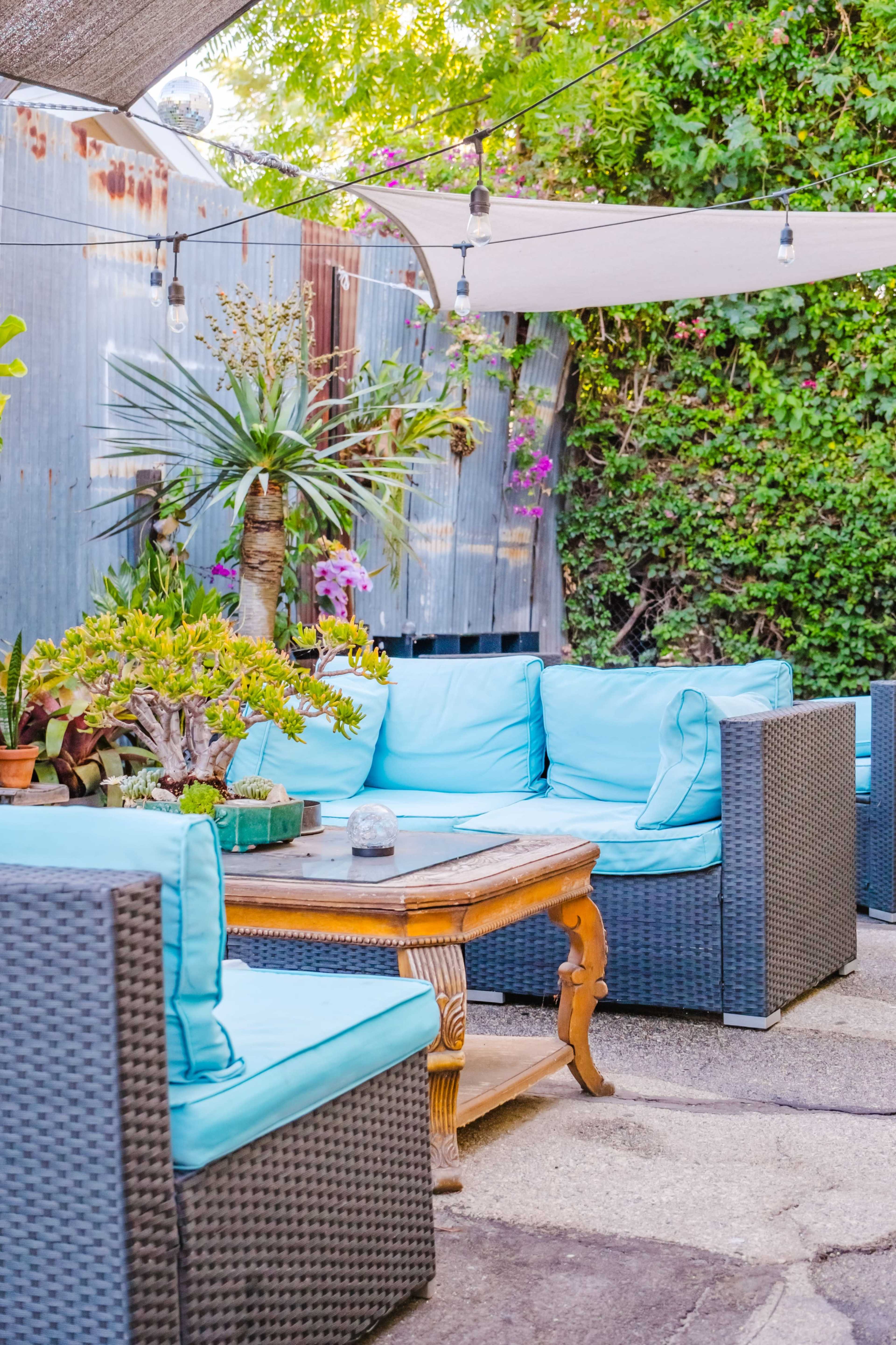 The image depicts a cozy outdoor seating area with turquoise cushions on woven furniture, a wooden coffee table, and surrounding greenery.
