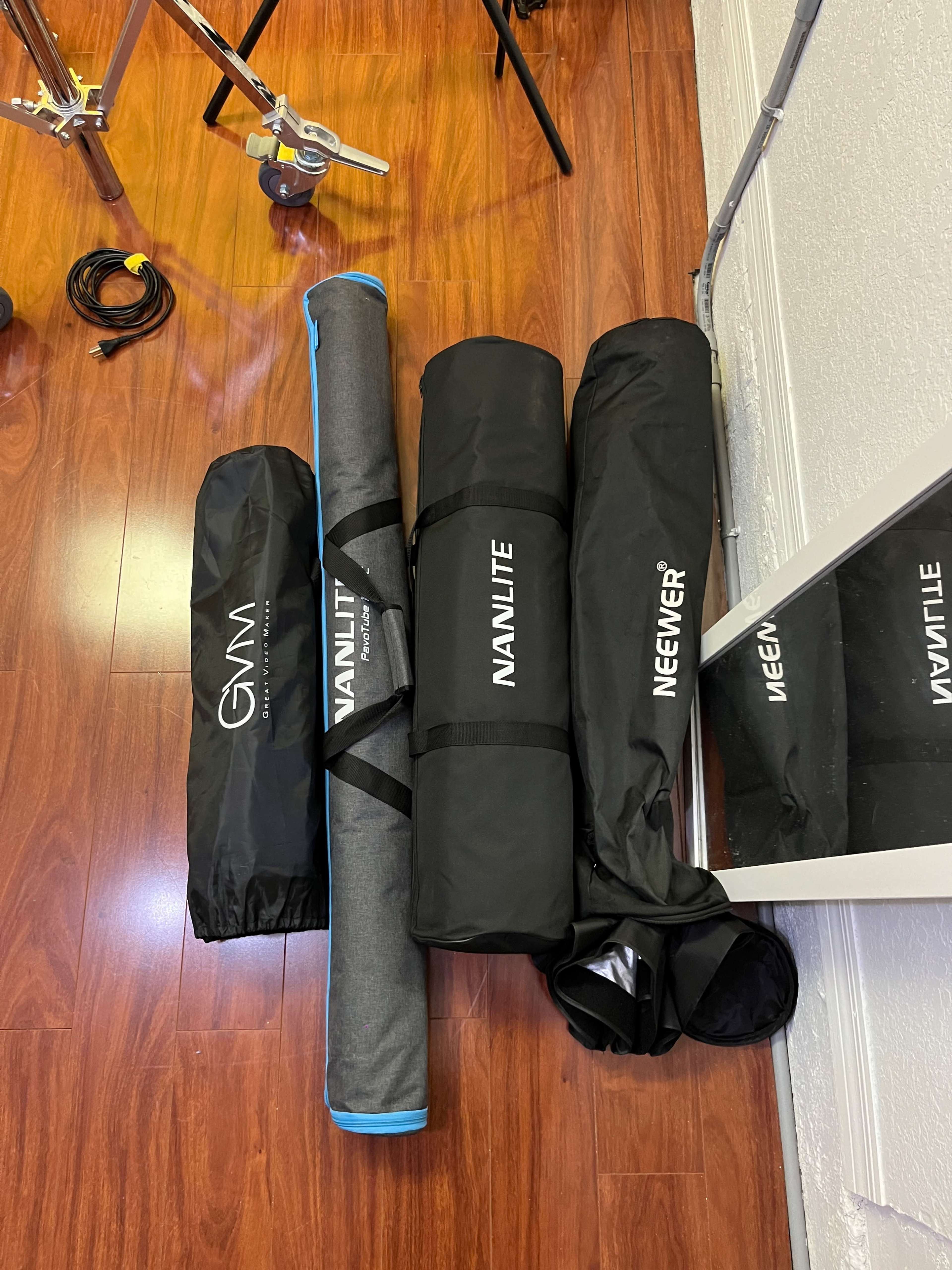 Several cylindrical equipment bags on a wooden floor, including brands like GVM, Nanlite, and Neewer.