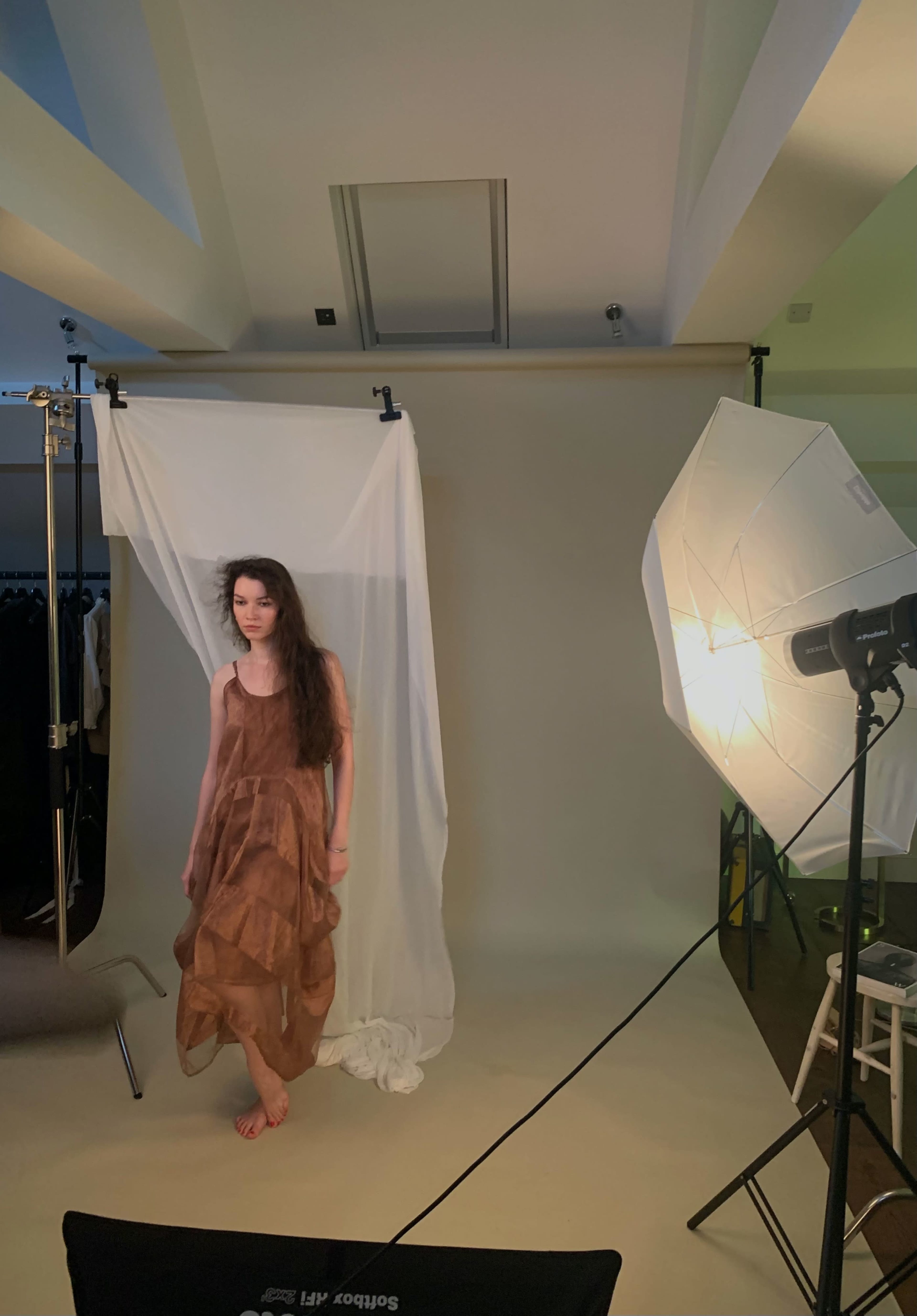 A model stands in a photography studio with a draped background and studio lighting equipment.