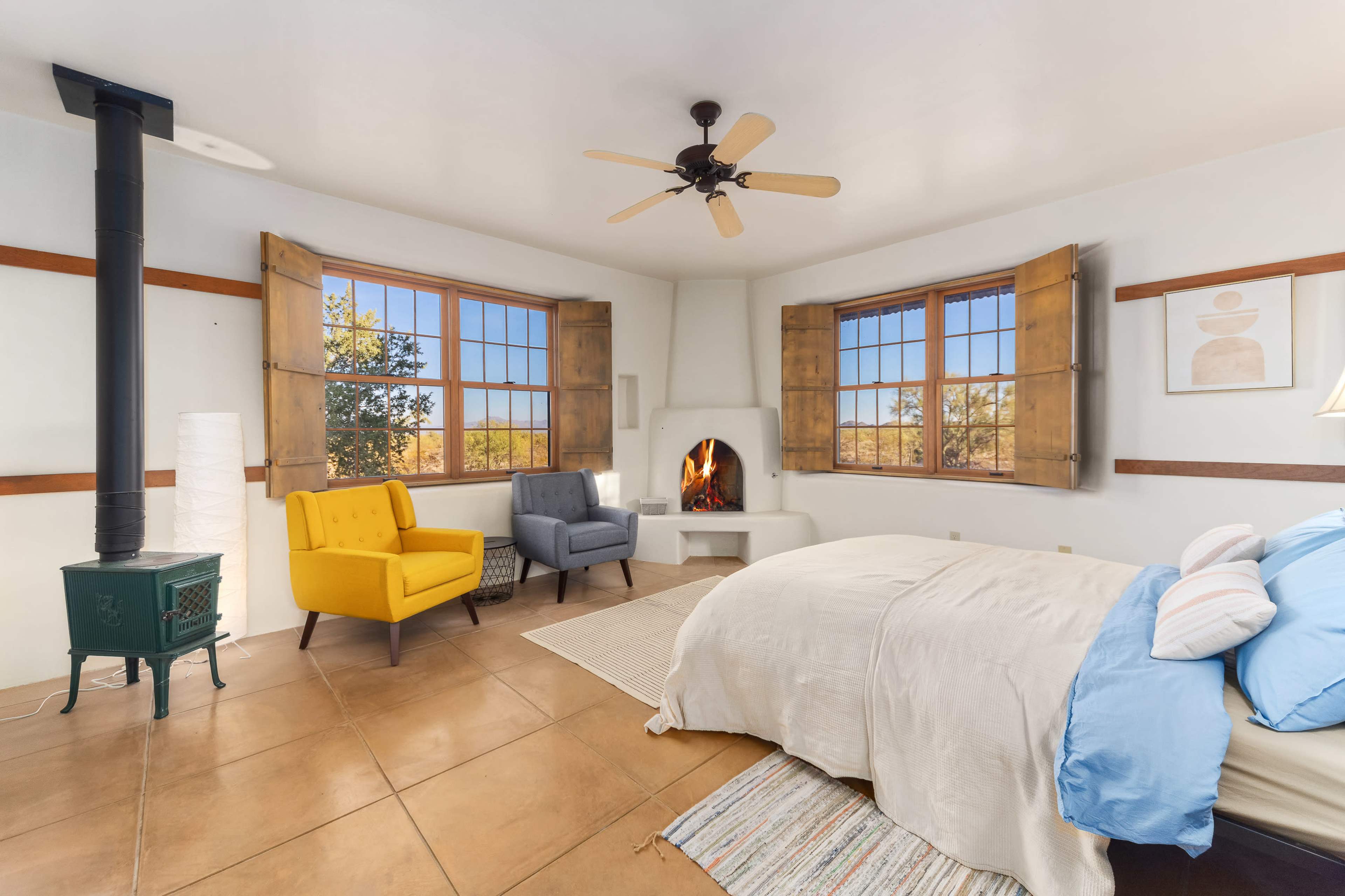 A bright bedroom features a bed with light bedding, two armchairs in yellow and gray, and a fireplace, with large windows offering views of the outdoors.