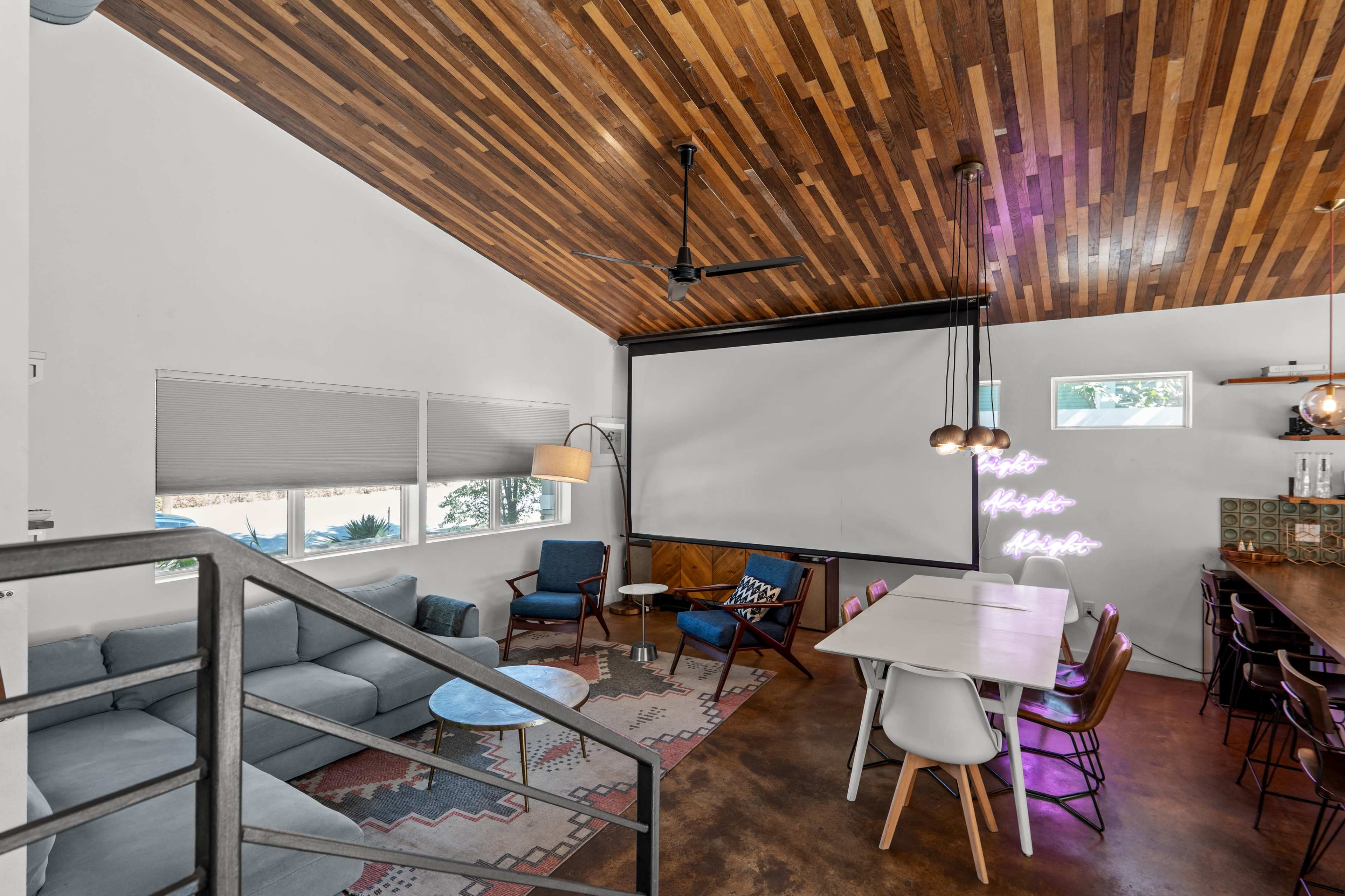 The image features a modern interior space with a wooden ceiling, a mix of seating options, and a large screen for presentations or movies.