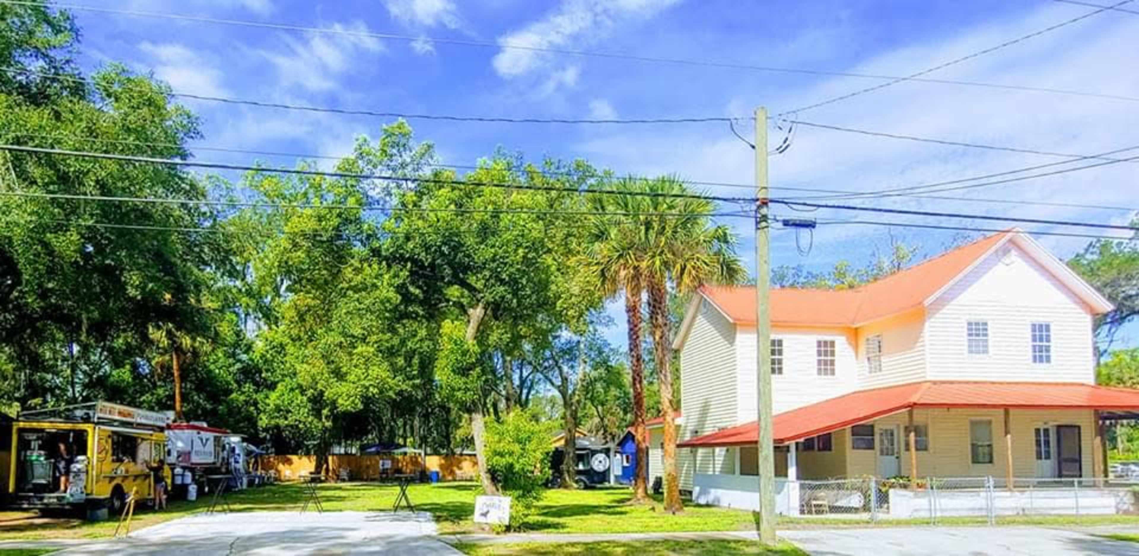 A white two-story house with a red roof is situated on a tree-lined street alongside a food truck and several large trees.
