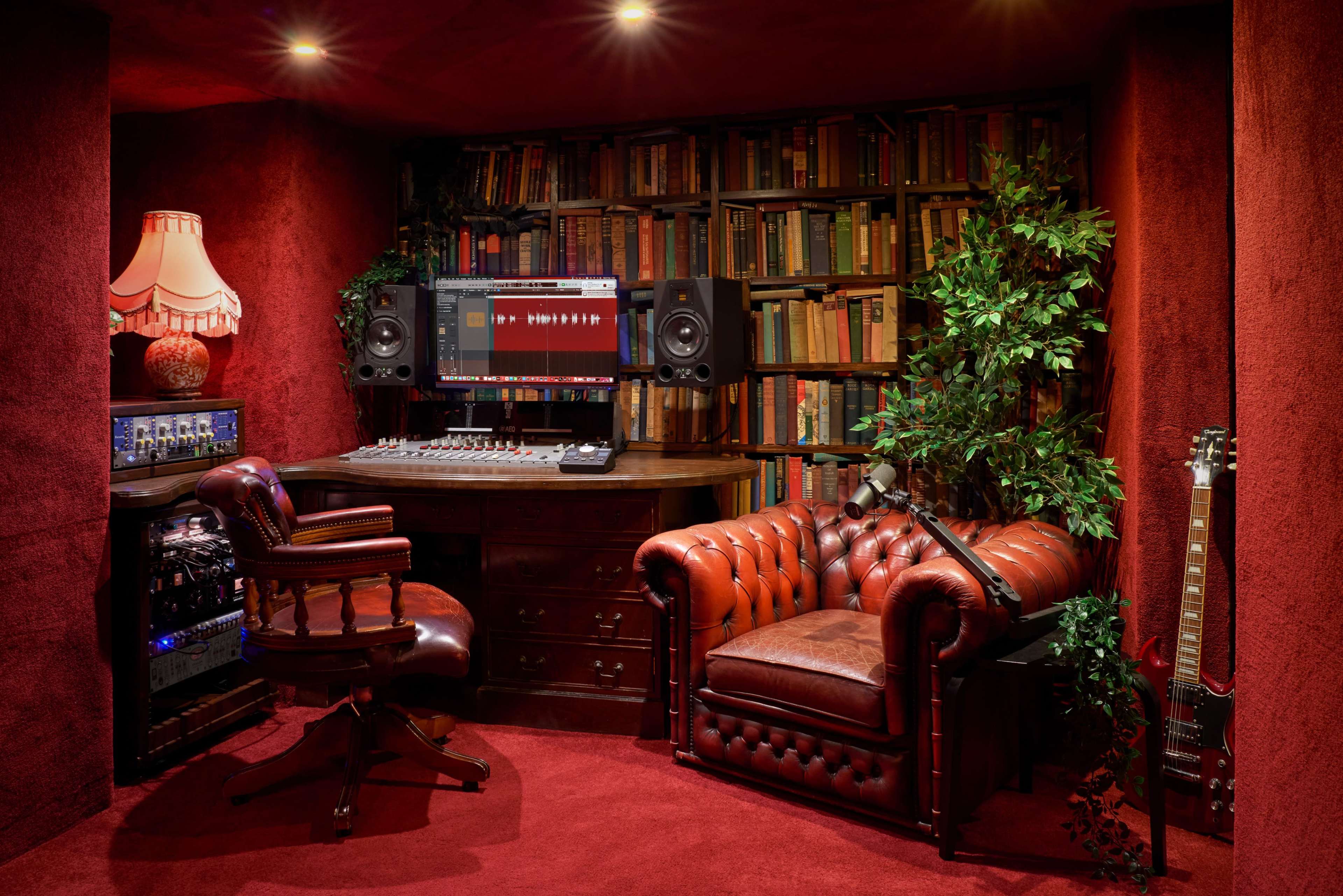 The image shows a recording studio with a wooden desk, audio equipment, a brown leather sofa, a library of books, and green plants.