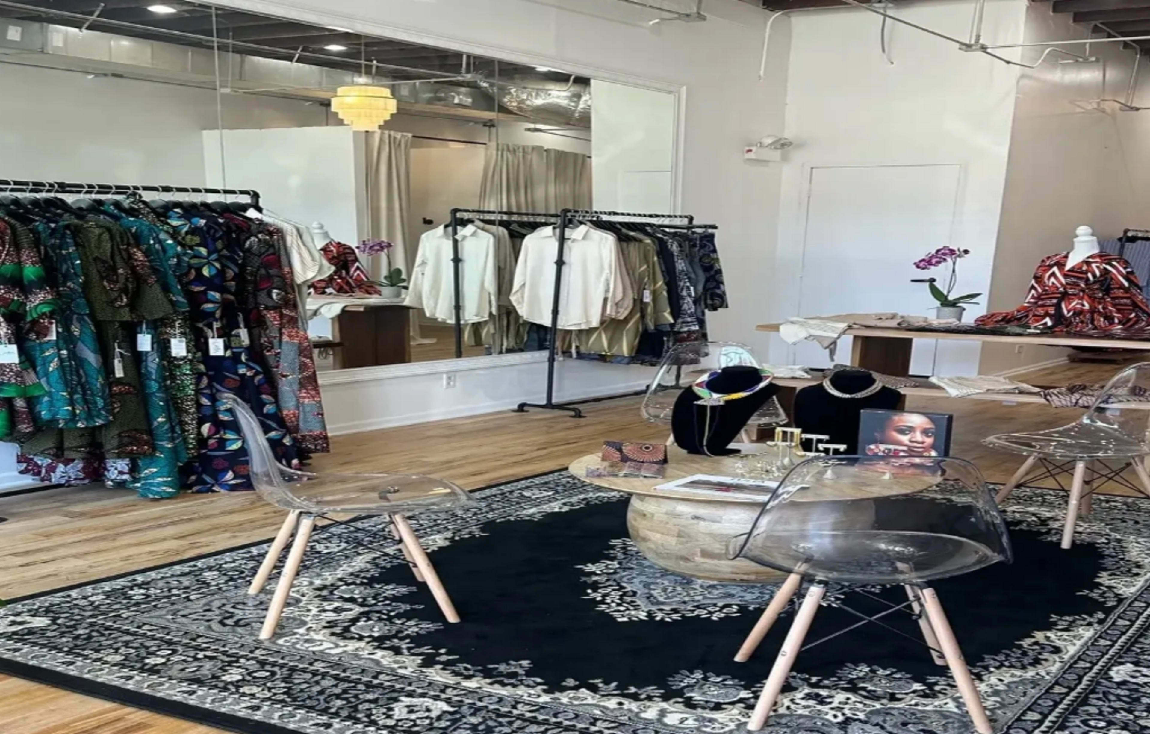 The image shows a boutique interior featuring clothing racks filled with various garments, a round table with accessories, and clear chairs arranged on a patterned rug.
