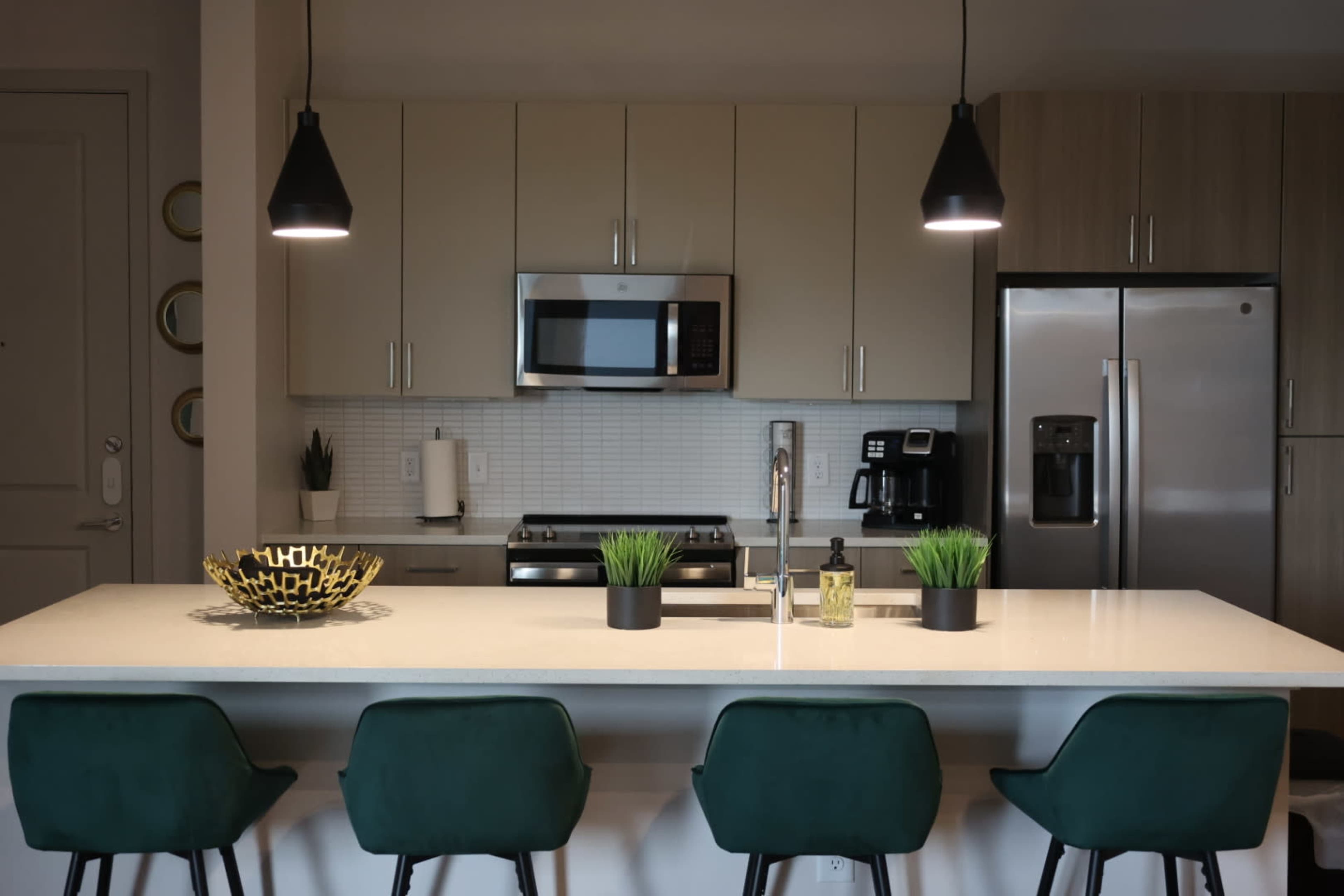A modern kitchen features a central island with four green bar stools, pendant lighting above, and stainless steel appliances.