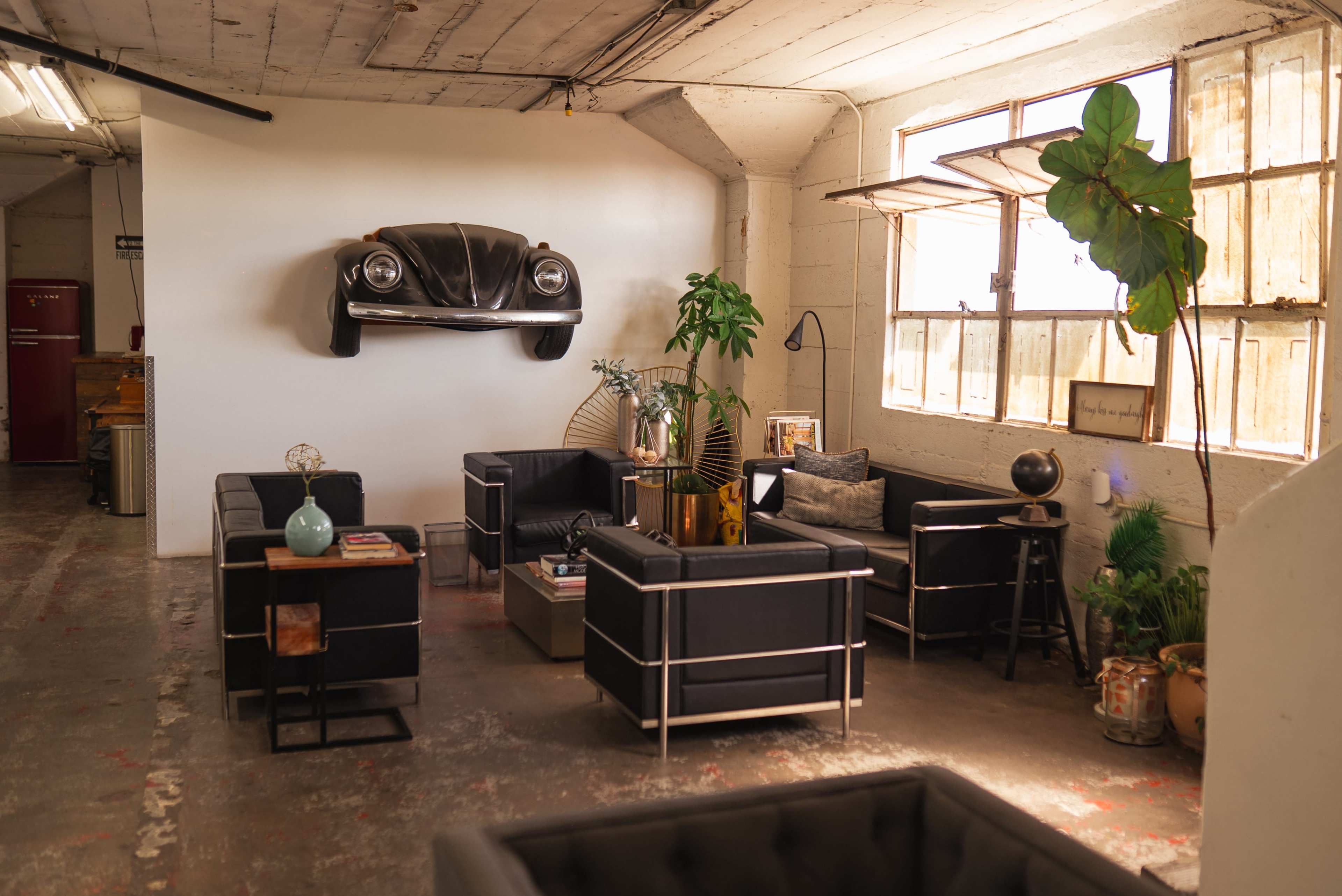 The image shows a modern lounge area with black furniture, a wall-mounted vintage car, and large windows allowing natural light to fill the space.