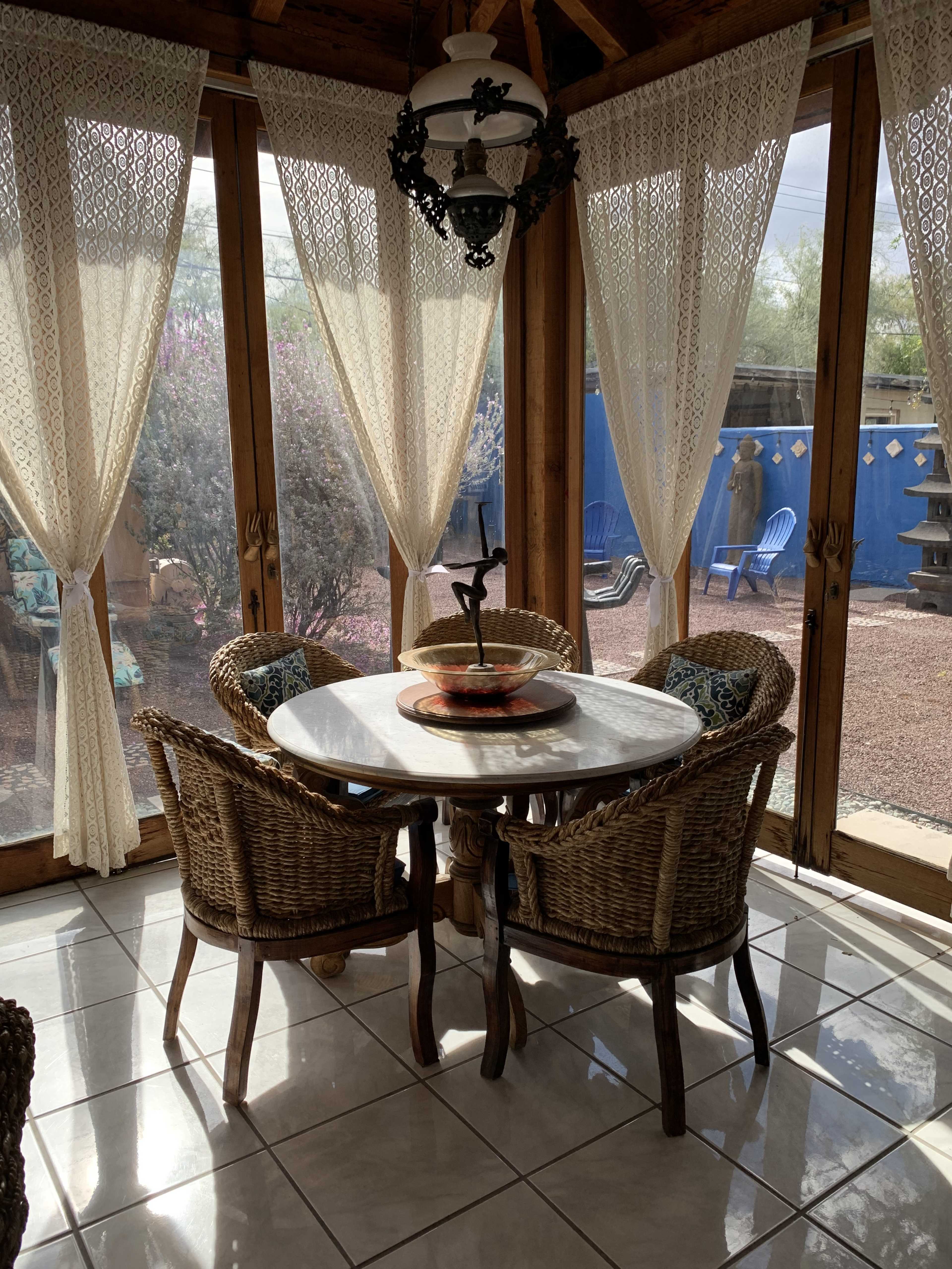 A round table with four wicker chairs is set in a sunlit room with lace curtains, overlooking a garden area with outdoor seating.