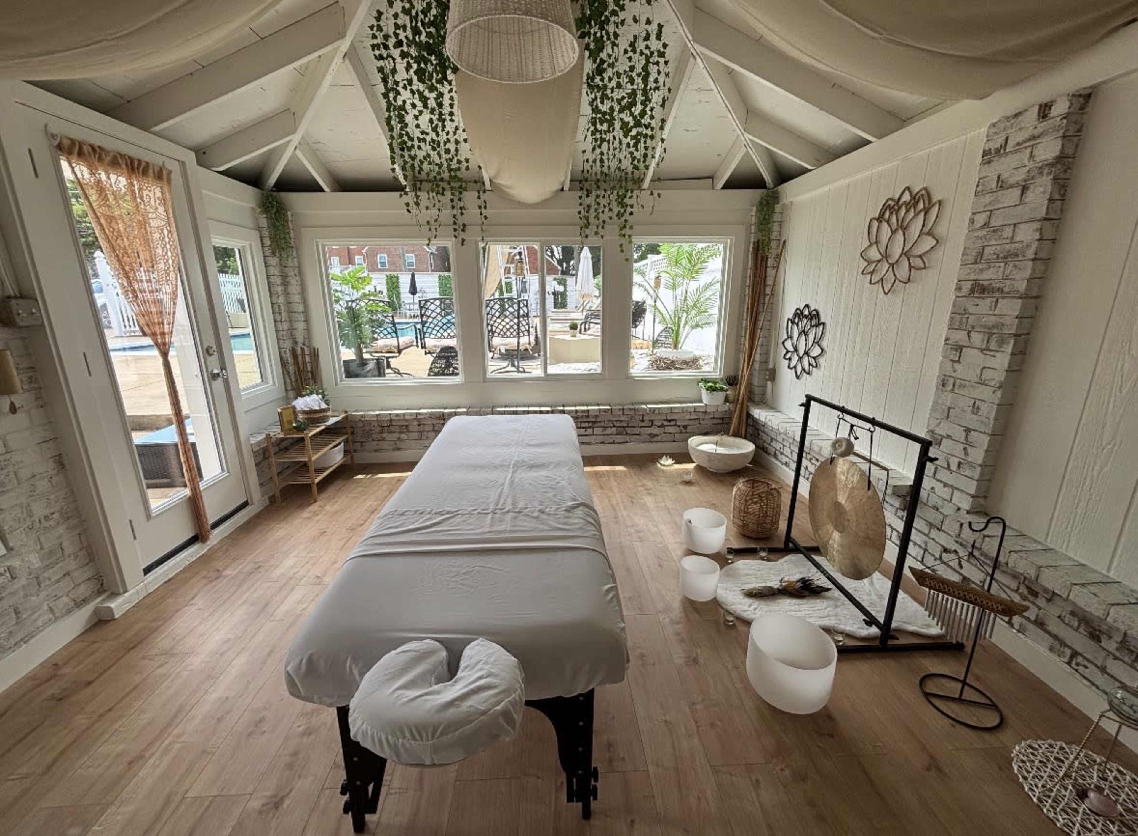 The image shows a serene massage room featuring a treatment table, decorative plants, and large windows that overlook an outdoor area with seating.