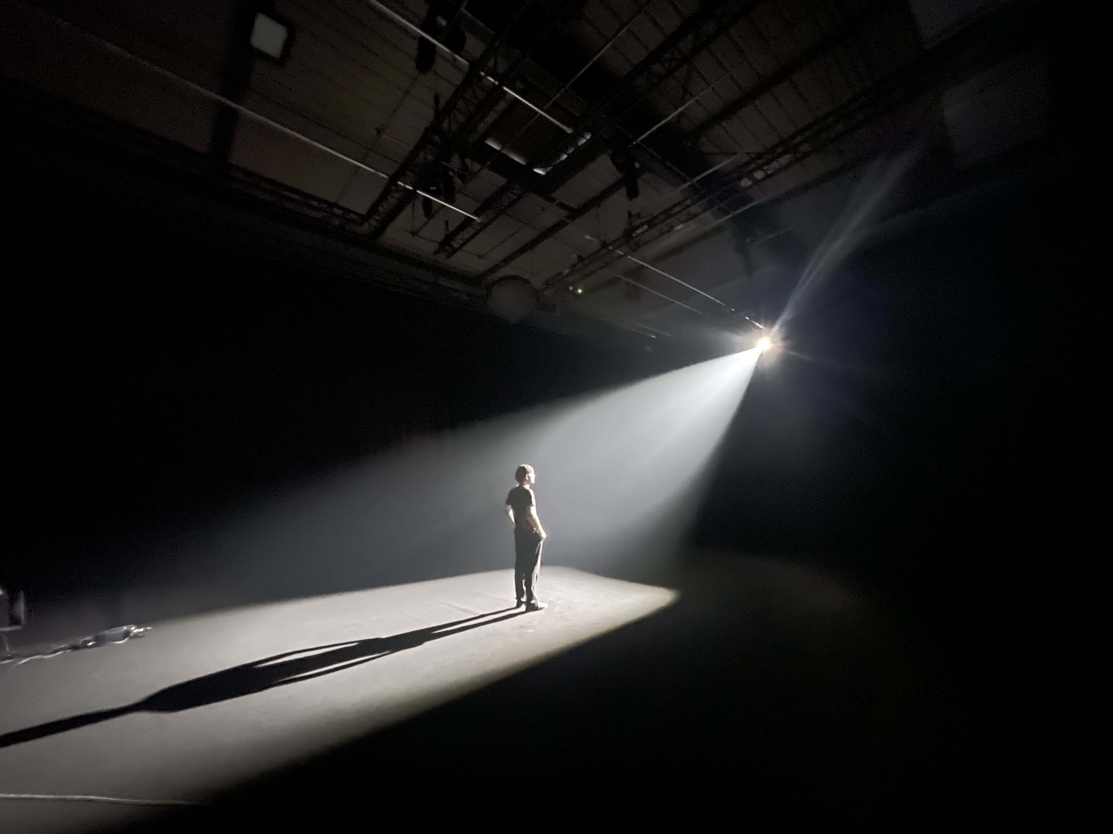 A single figure stands on a dimly lit stage, illuminated by a stark beam of light from above.
