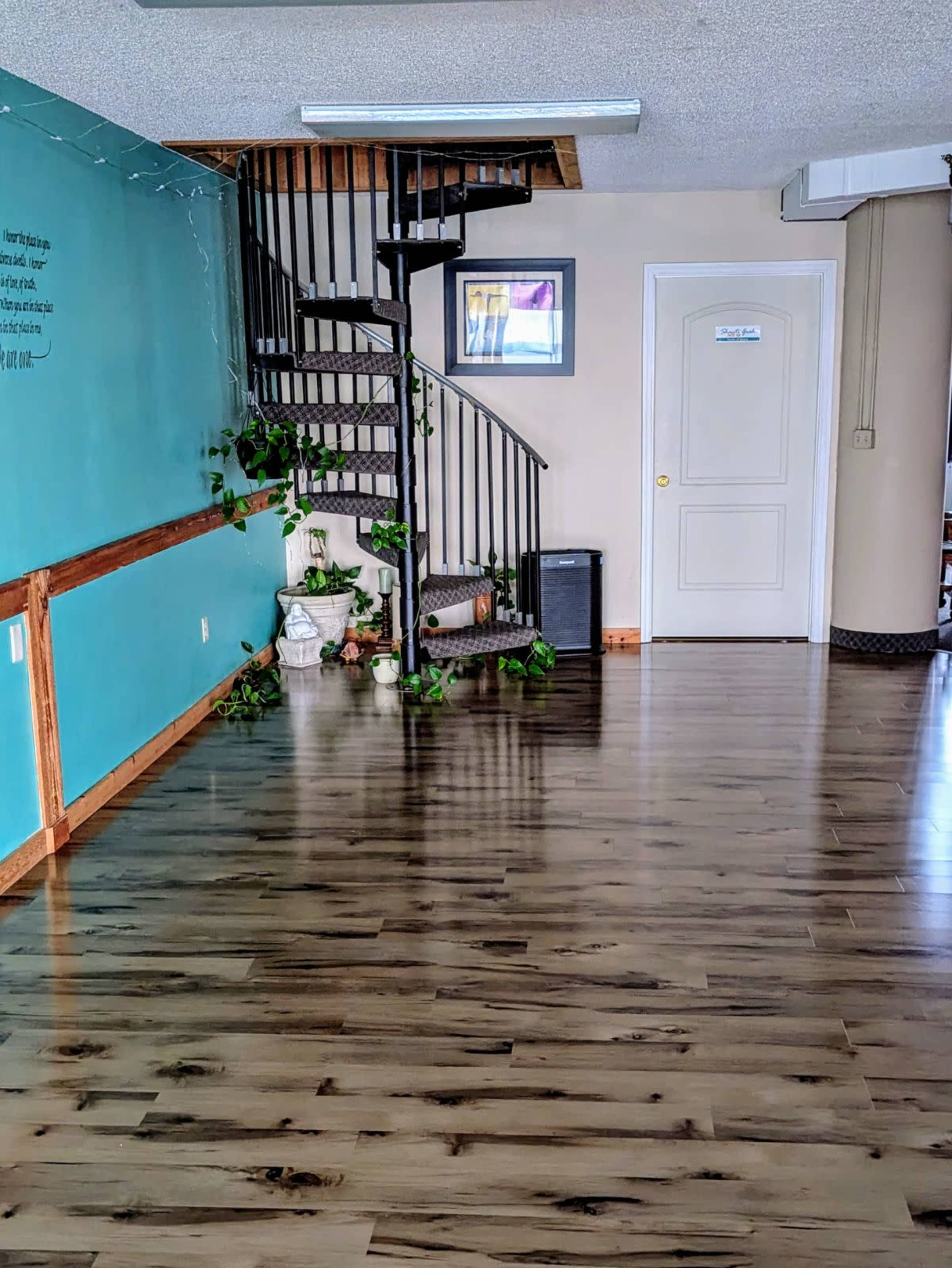 The image shows a spacious interior with a wooden floor, a spiral staircase leading to an upper level, and a wall painted in turquoise adorned with plants and a framed picture.
