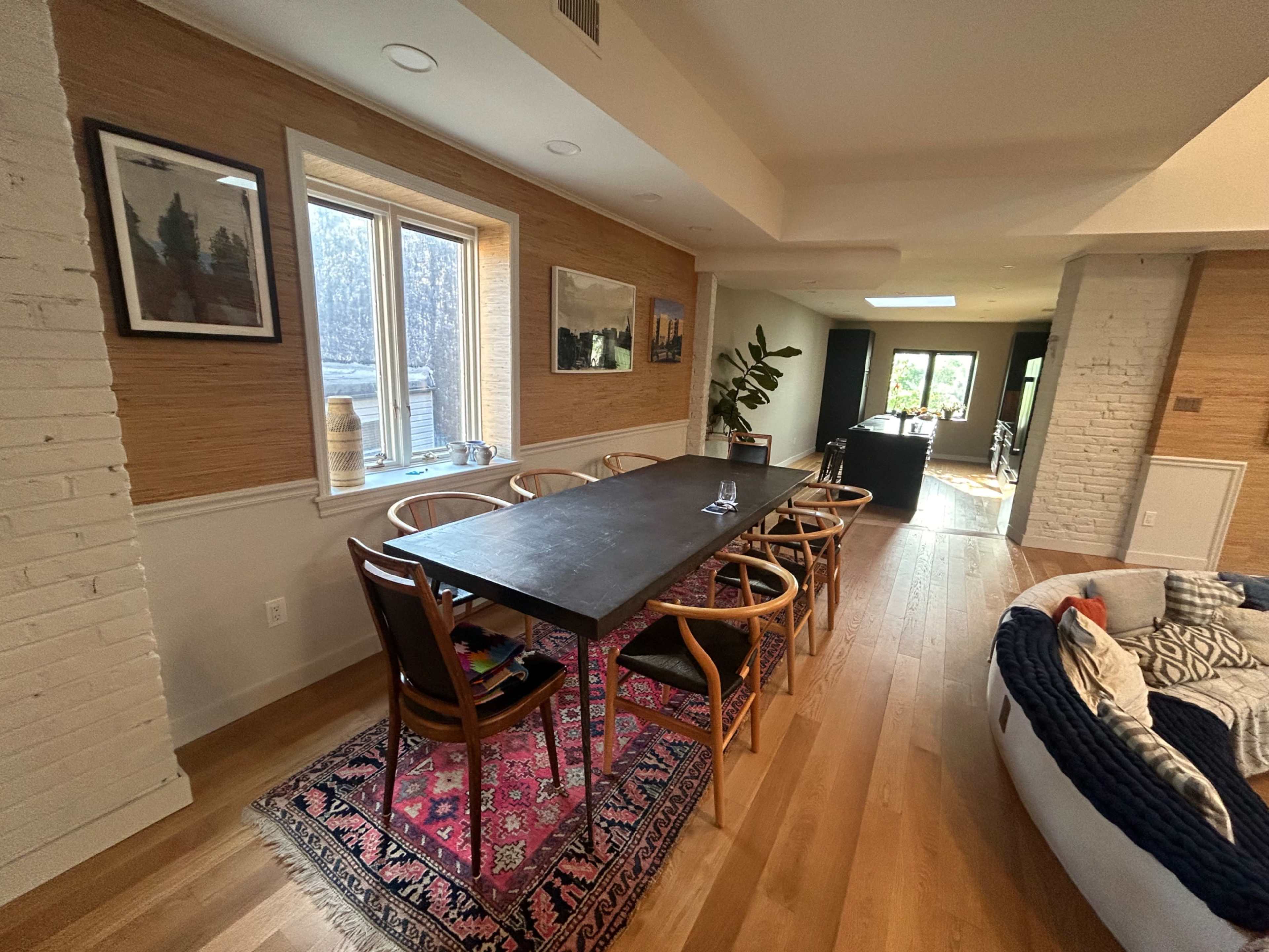 A spacious dining area features a long dark wooden table surrounded by wooden chairs, with a colorful rug underneath and large windows letting in natural light.
