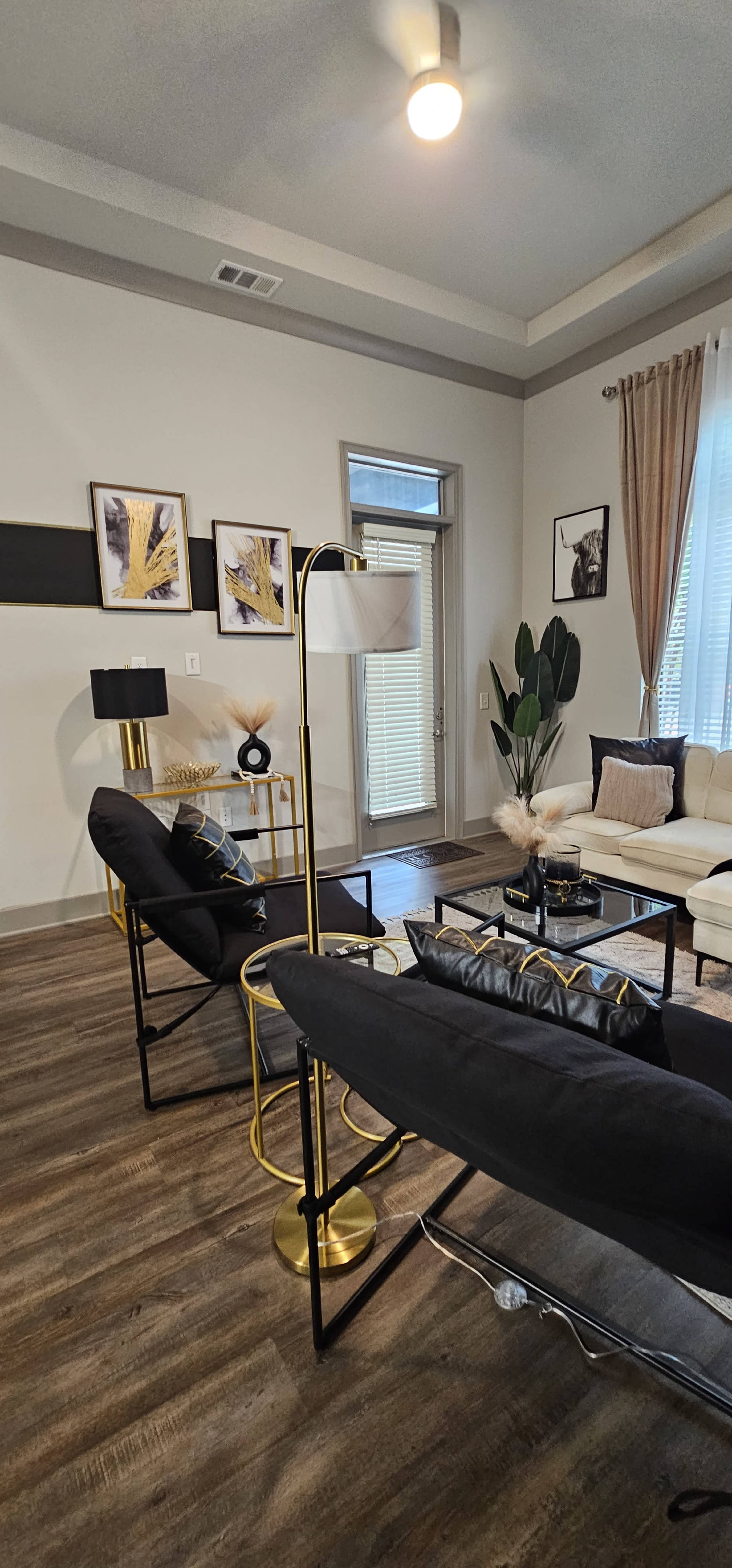 The image shows a modern living room with two black chairs, a cream sofa, decorative wall art, and a floor lamp, all arranged around a coffee table.