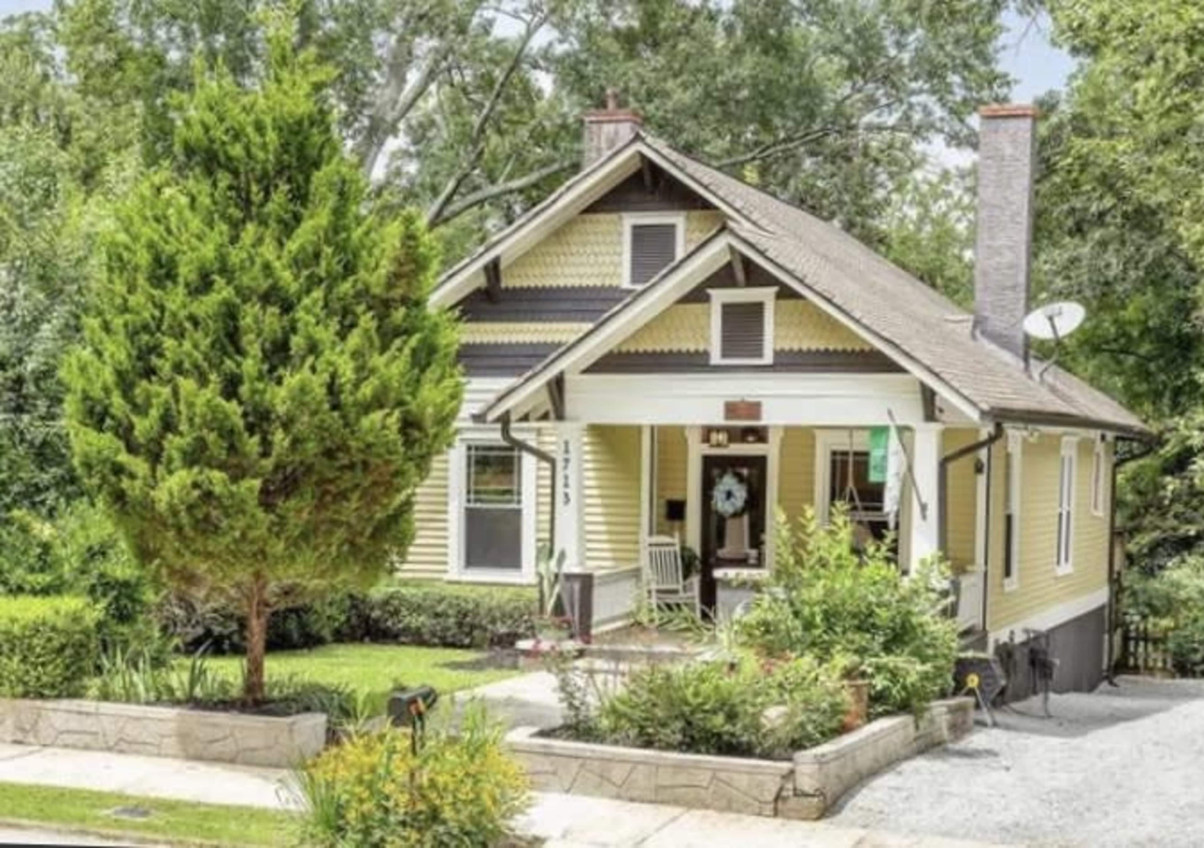 A yellow bungalow with a front porch is surrounded by landscaped greenery and a gravel path.