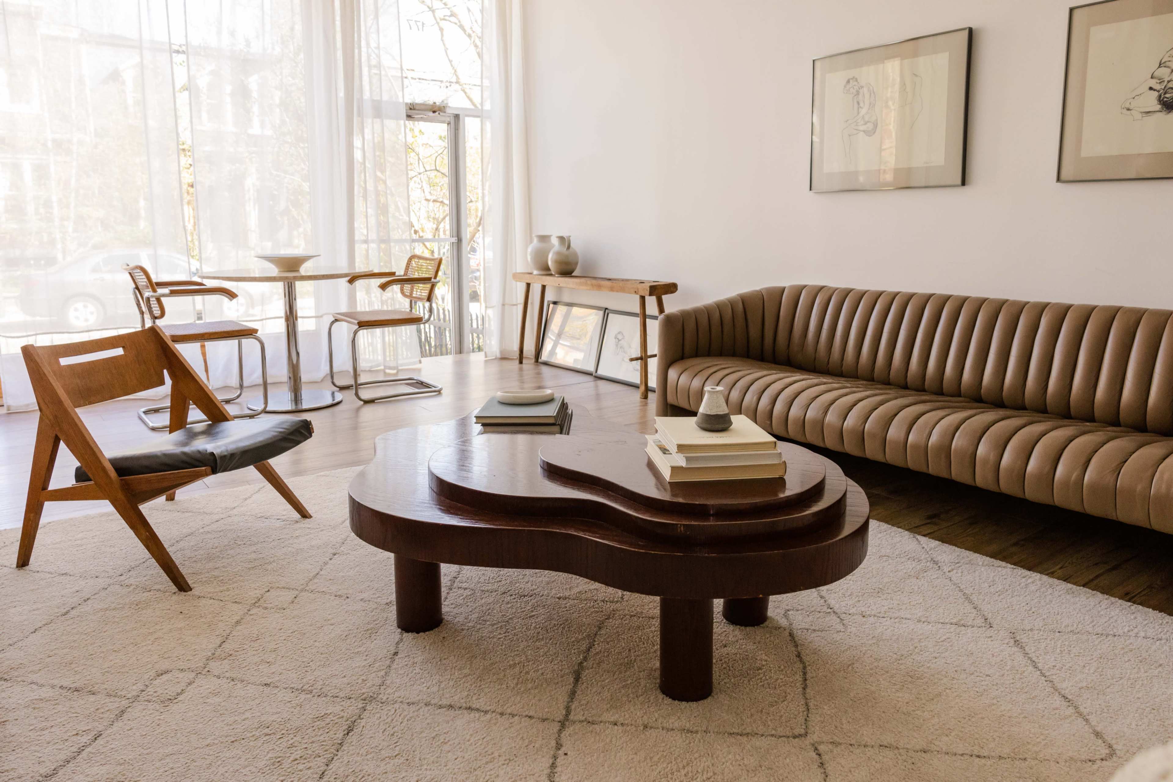A minimalist living room featuring a curved brown sofa, a wooden coffee table, a patterned rug, and a dining area with chairs and a table.