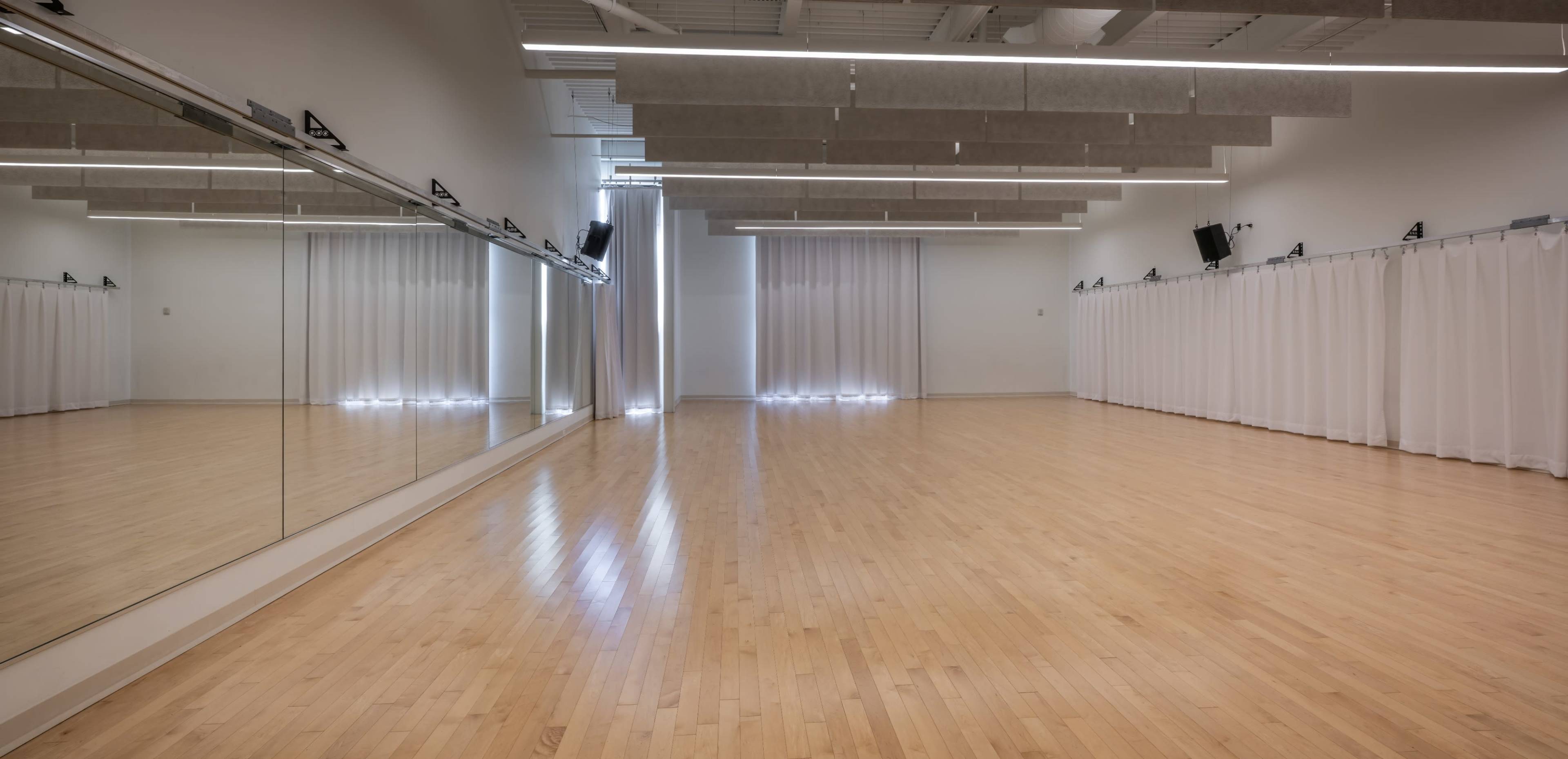 The image shows a spacious dance studio with wooden flooring, large mirrors, and white curtains.