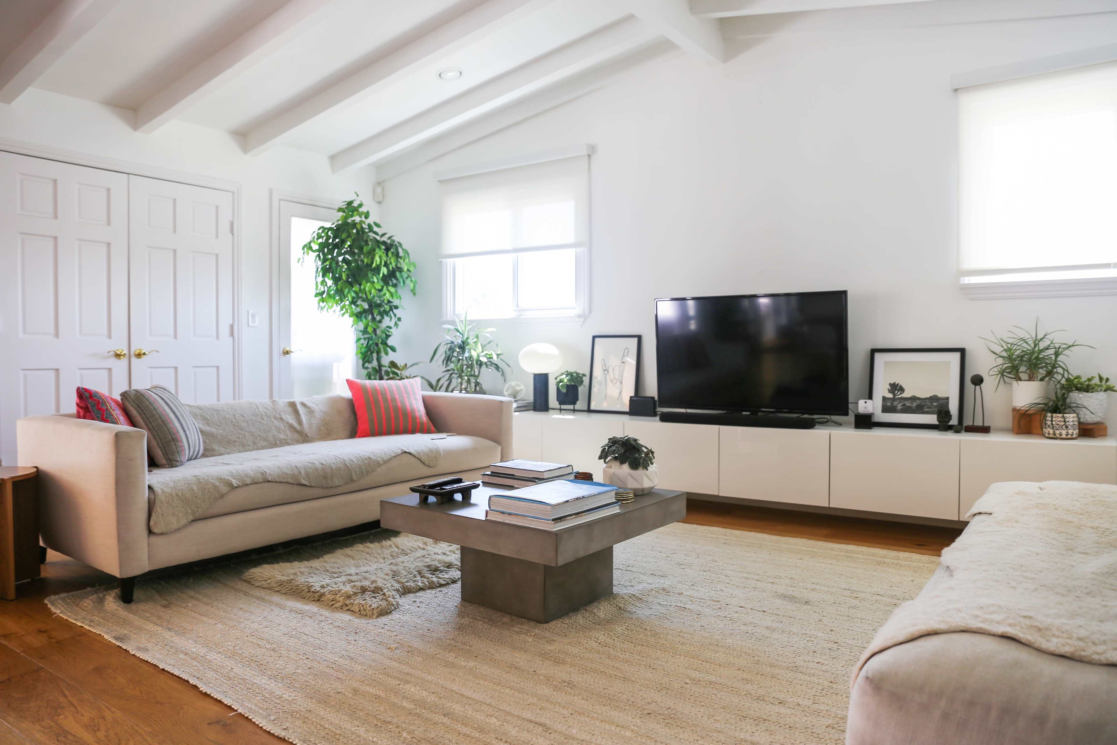 The room features a modern living space with a beige sofa, a low coffee table, a large television, and several potted plants, all set against a backdrop of white walls and wooden flooring.