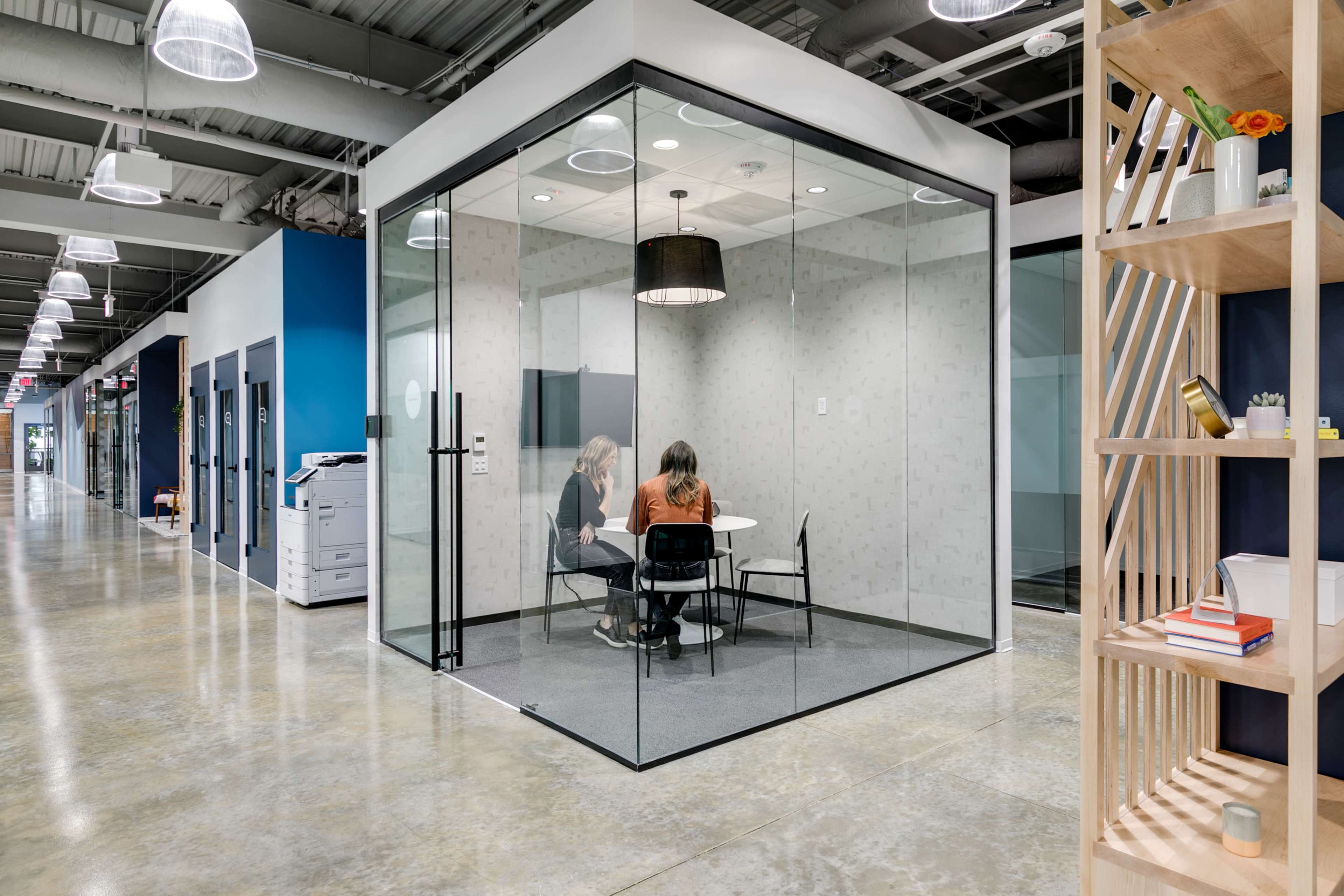 A glass-walled meeting room with two people seated at a table is located in a modern office corridor with blue and gray walls.