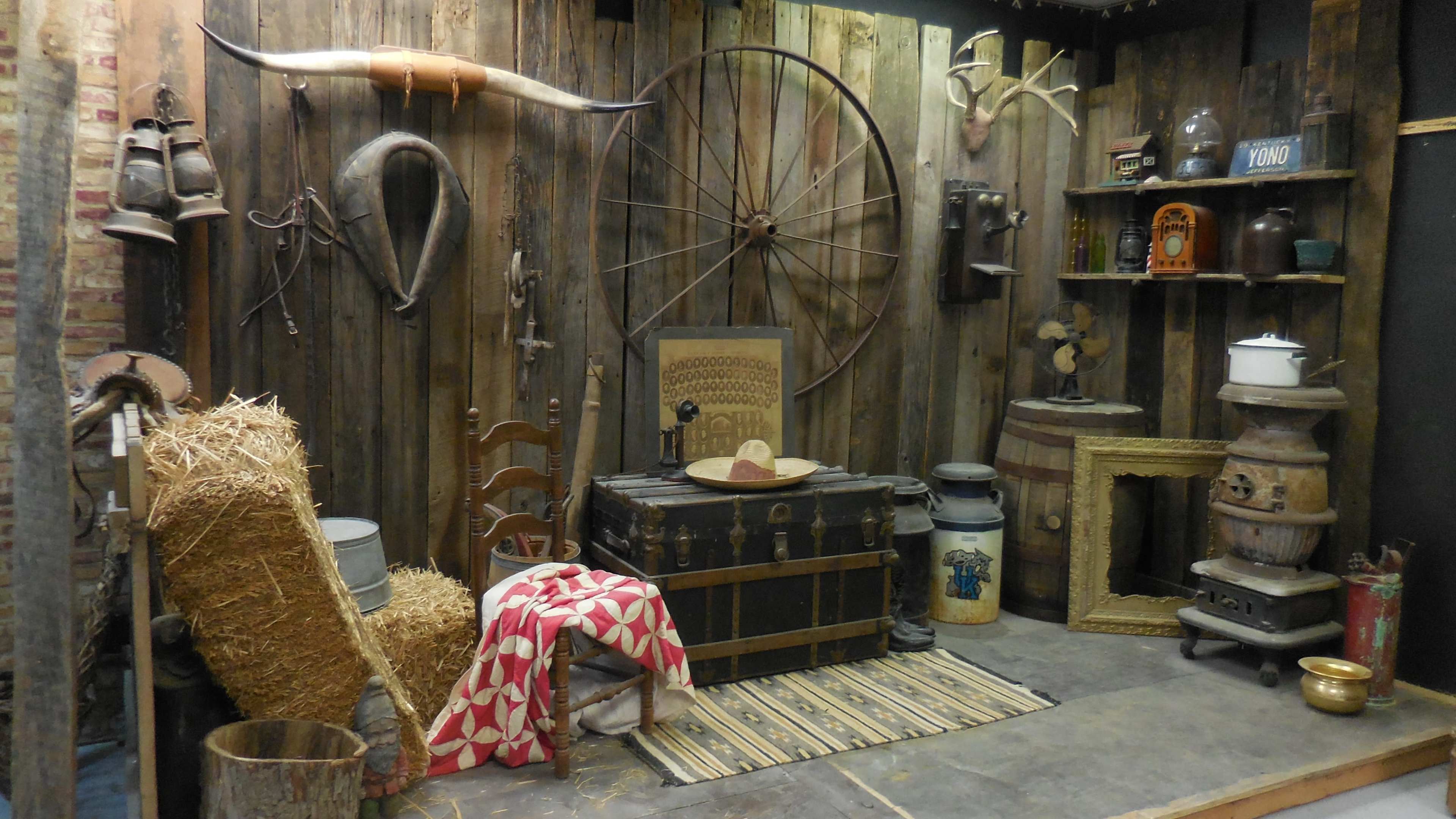 A rustic room decorated with wooden walls, various antique farm tools, a barrel, a chest, and some hay bales.