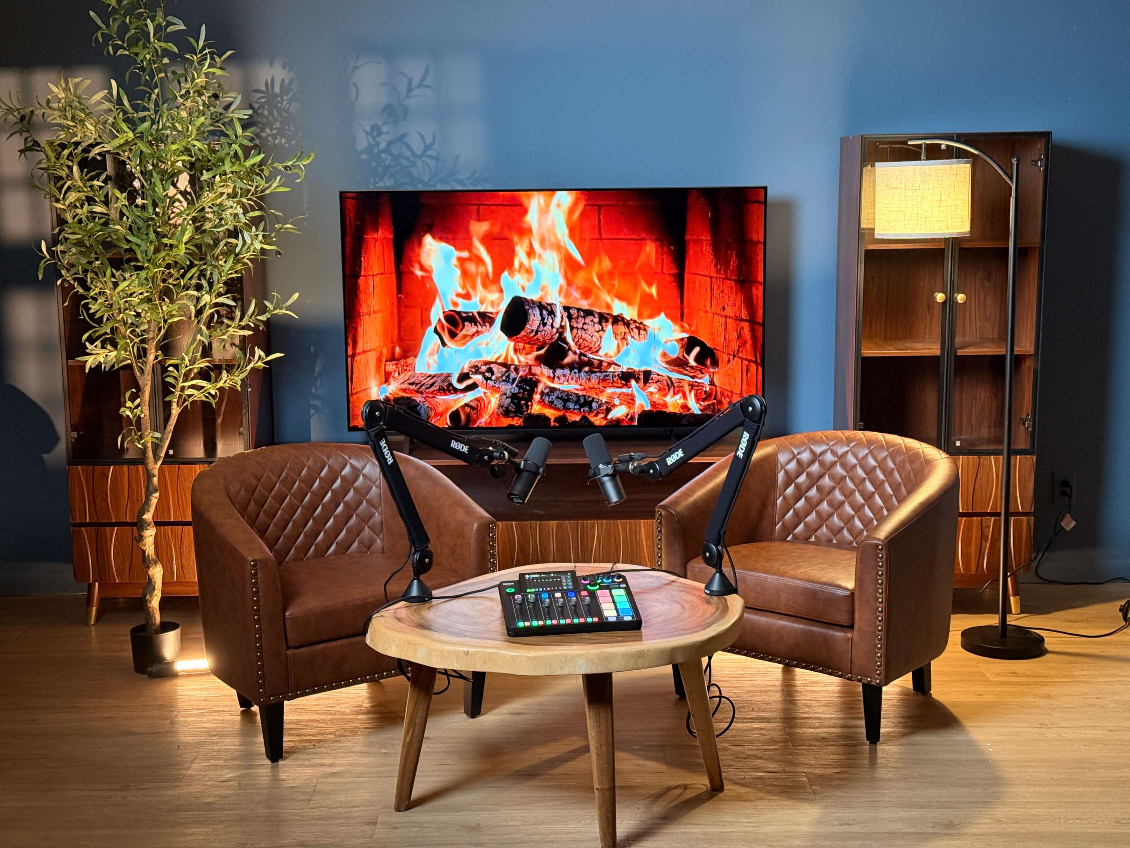 The image shows a cozy podcasting setup featuring two brown leather chairs, a round wooden table with a sound mixer, and a large television displaying a crackling fire in a blue room.