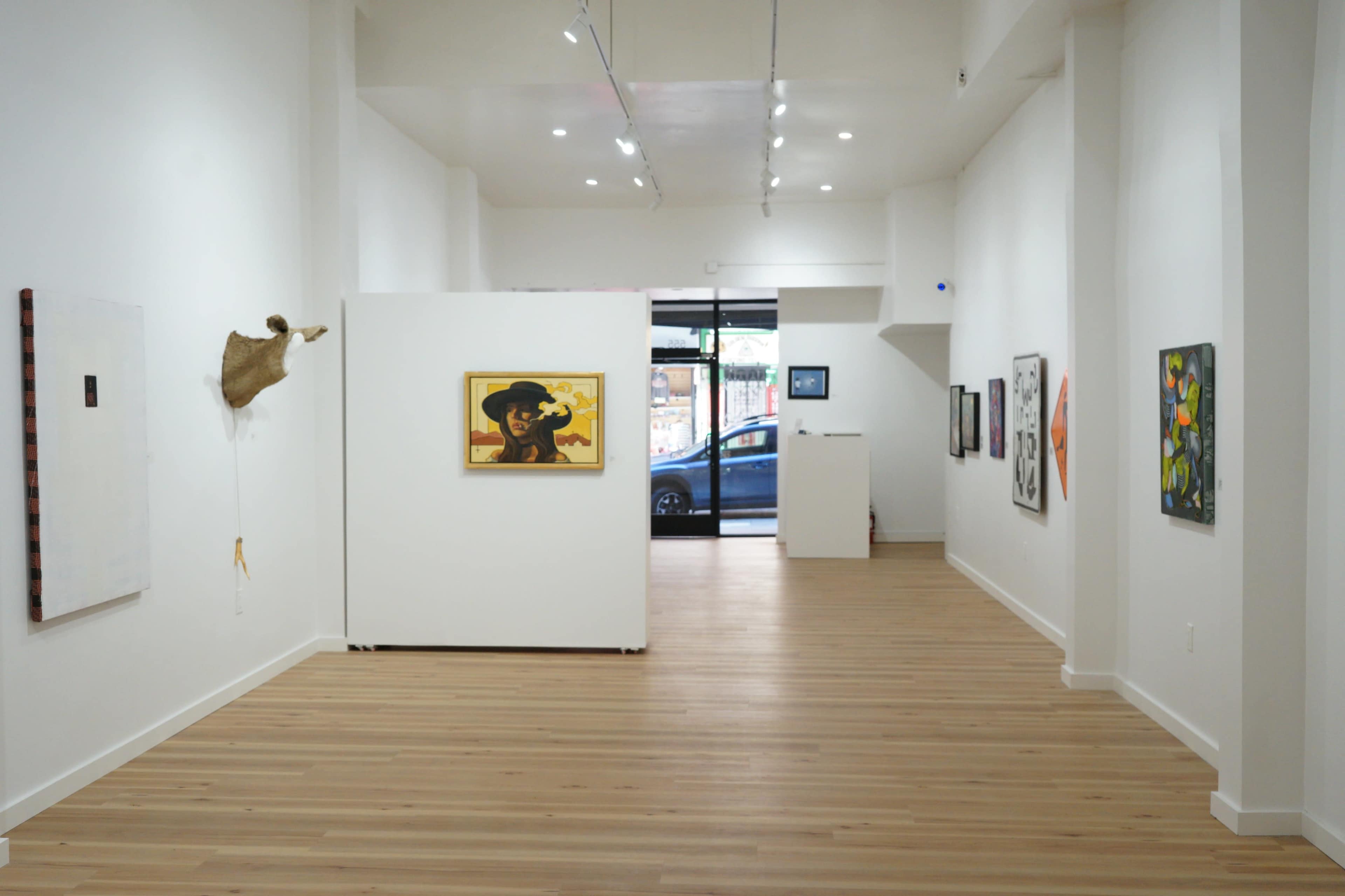 The interior of an art gallery features white walls, wooden flooring, and various artworks displayed, with a large window showing a car parked outside.