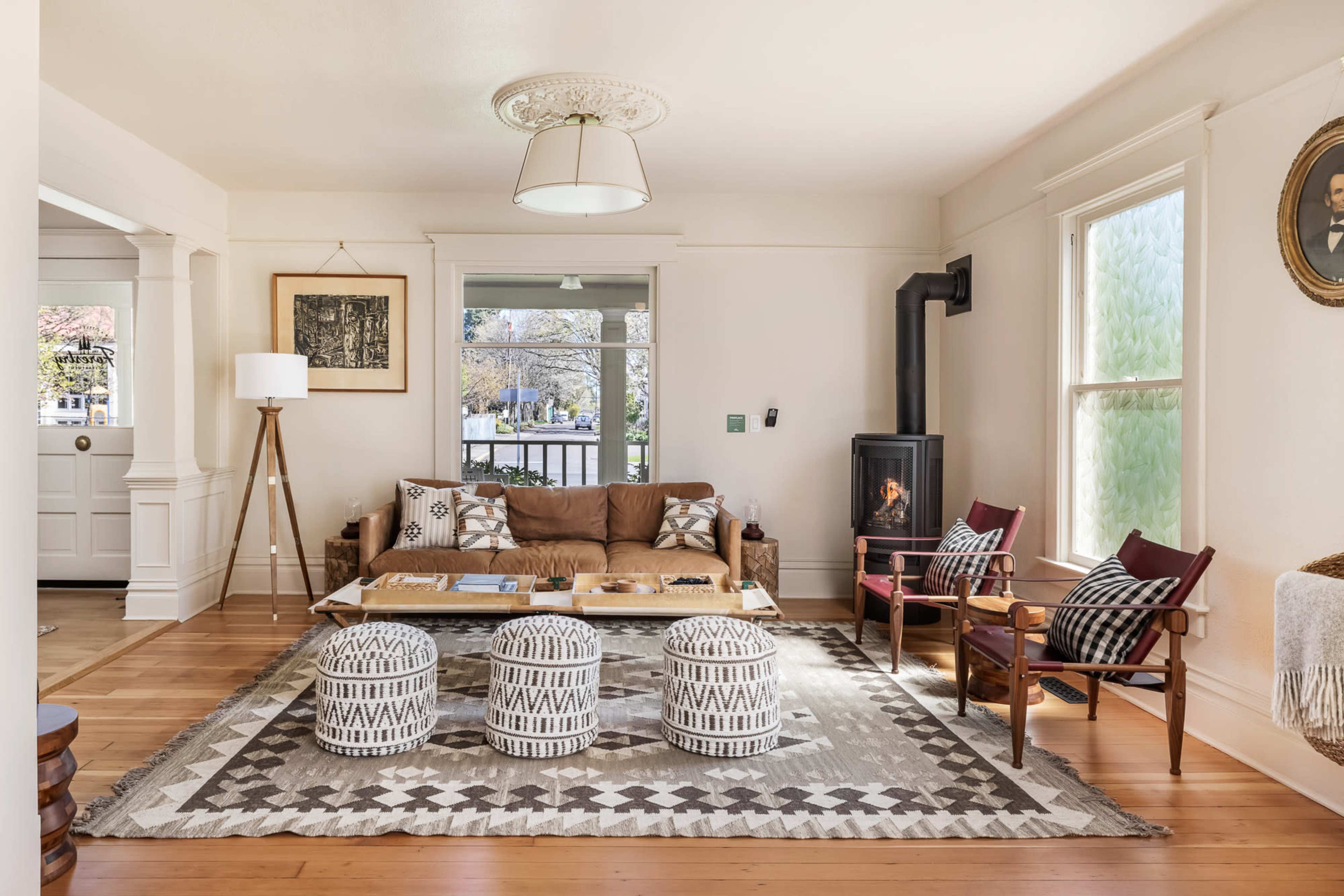 The image shows a cozy living room with a brown couch, a wooden coffee table, three patterned poufs, and a small wood stove beside large windows.
