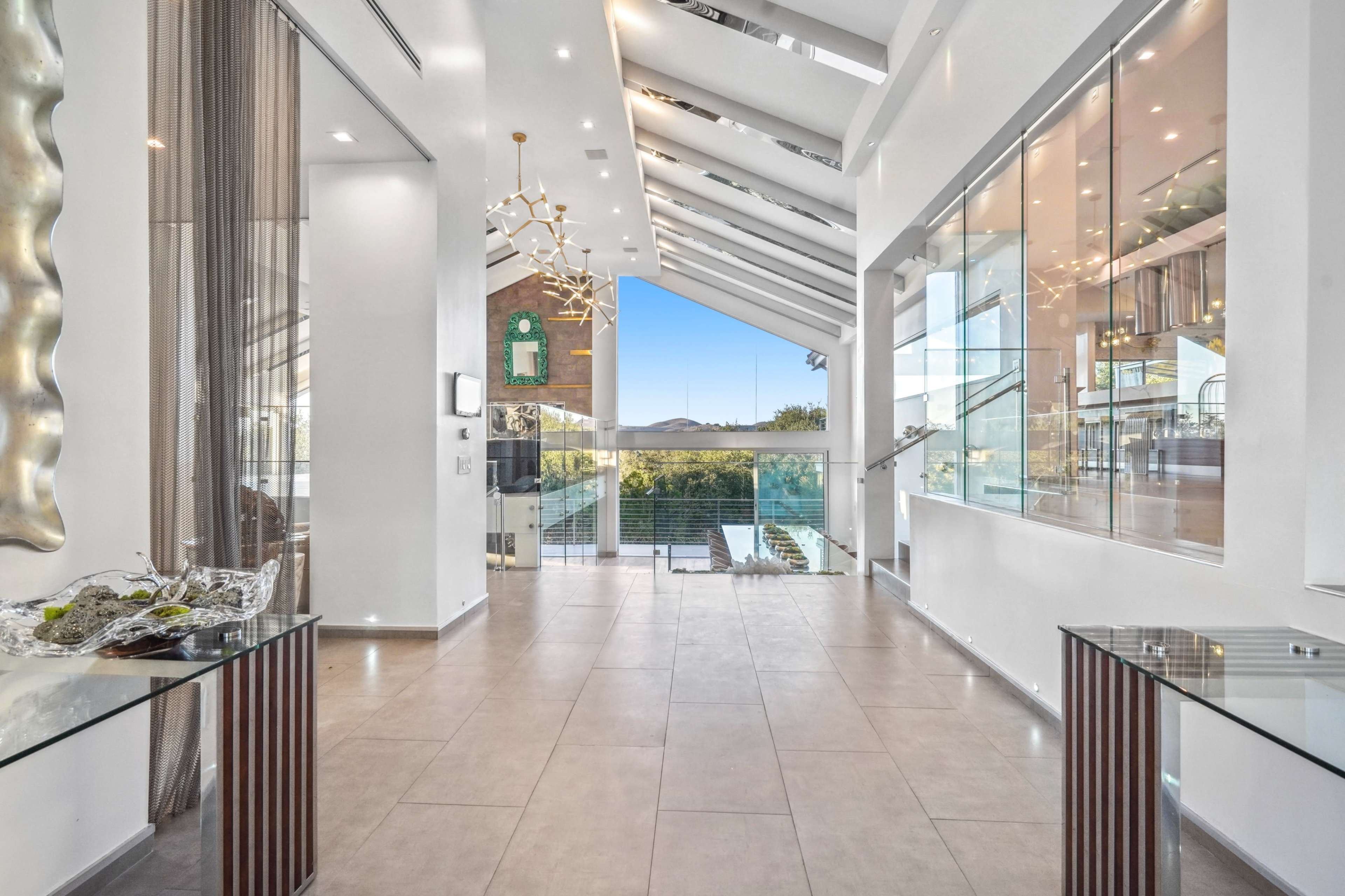 The image shows a modern interior with high ceilings, large glass windows, and a tiled floor, featuring a foyer leading to an outdoor space.