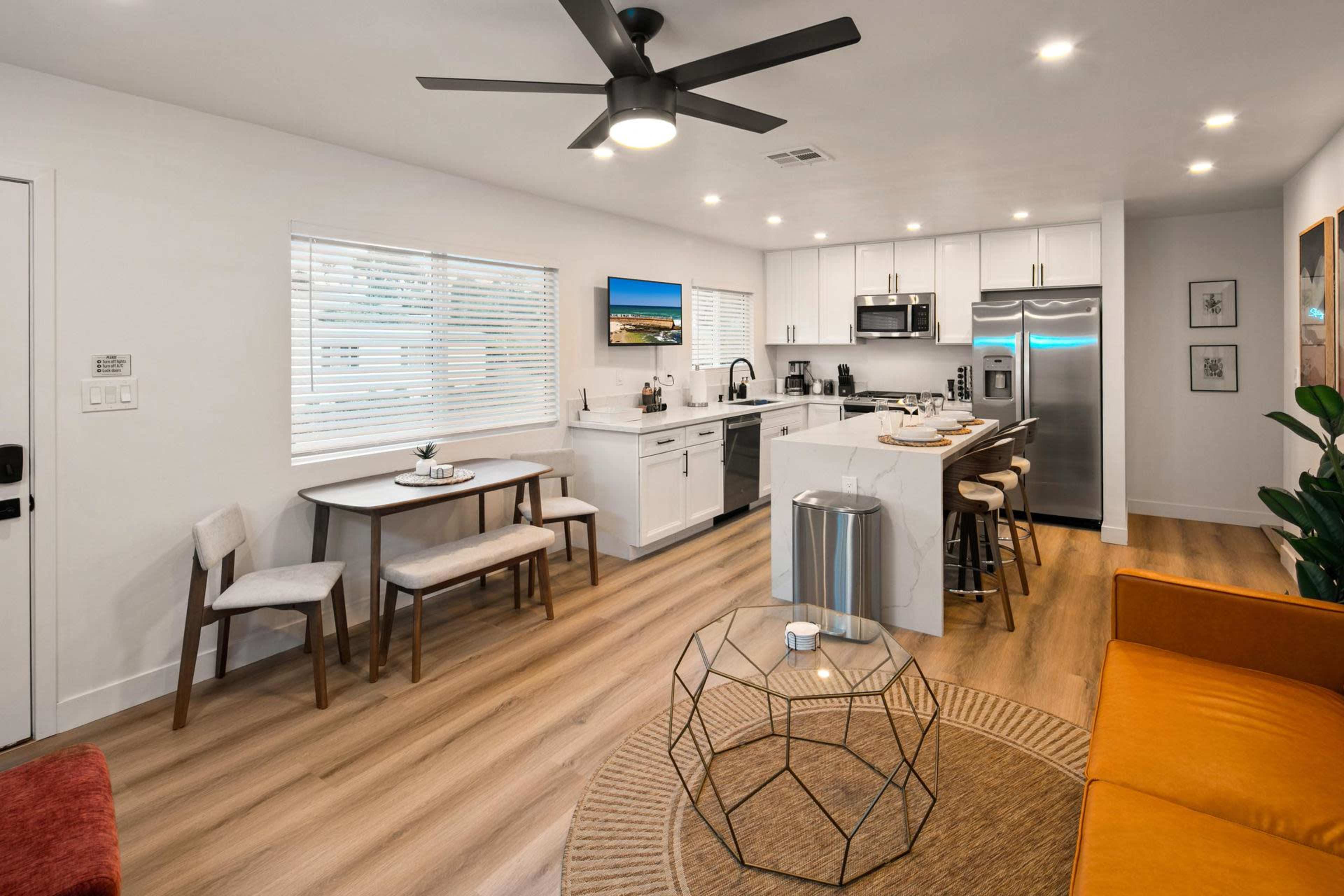 The image shows a modern, open-concept kitchen and living area with a dining table, a sofa, and large windows allowing natural light.