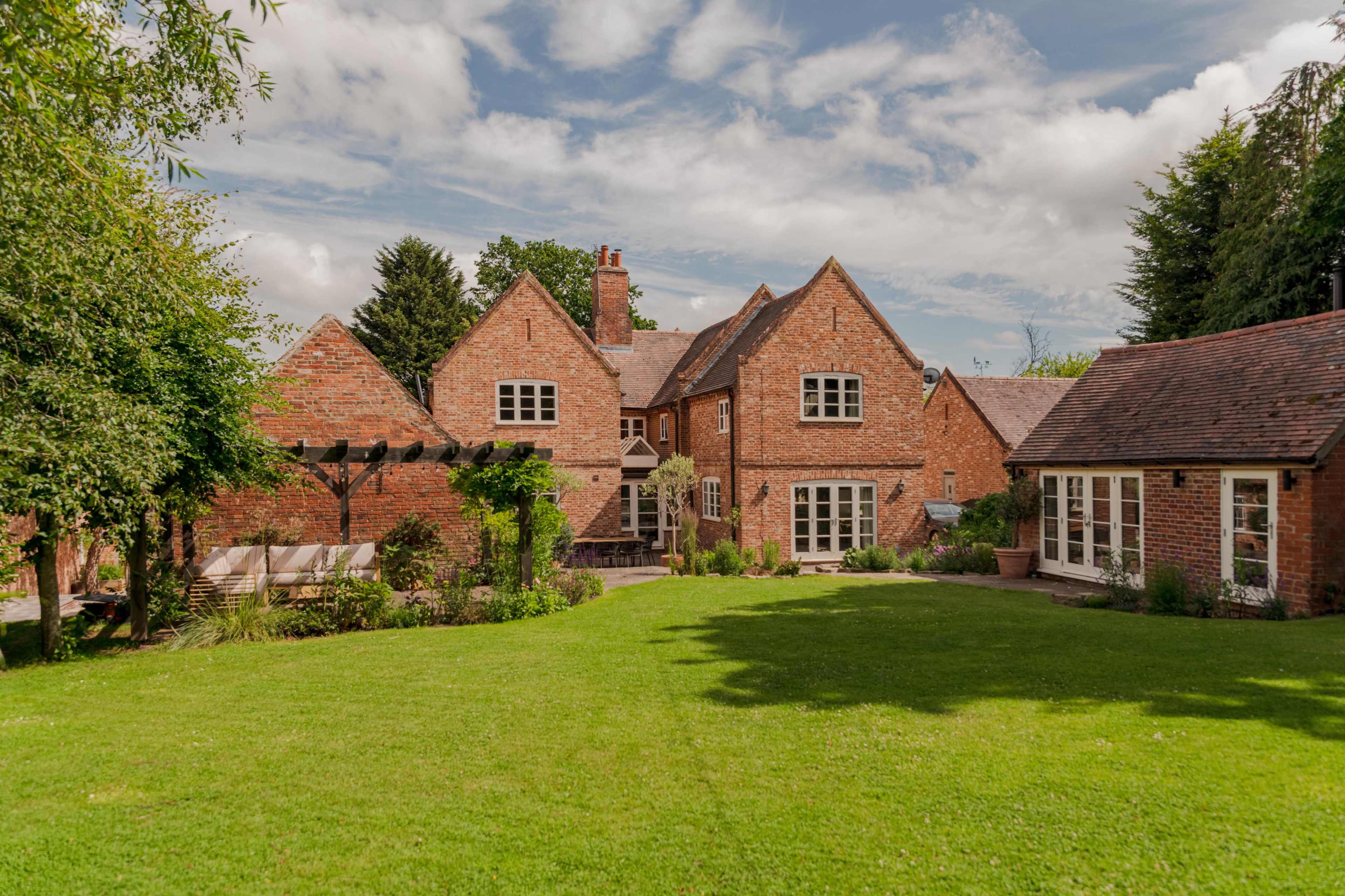 The image shows a brick house with multiple sections surrounded by a large green lawn and trees under a partly cloudy sky.