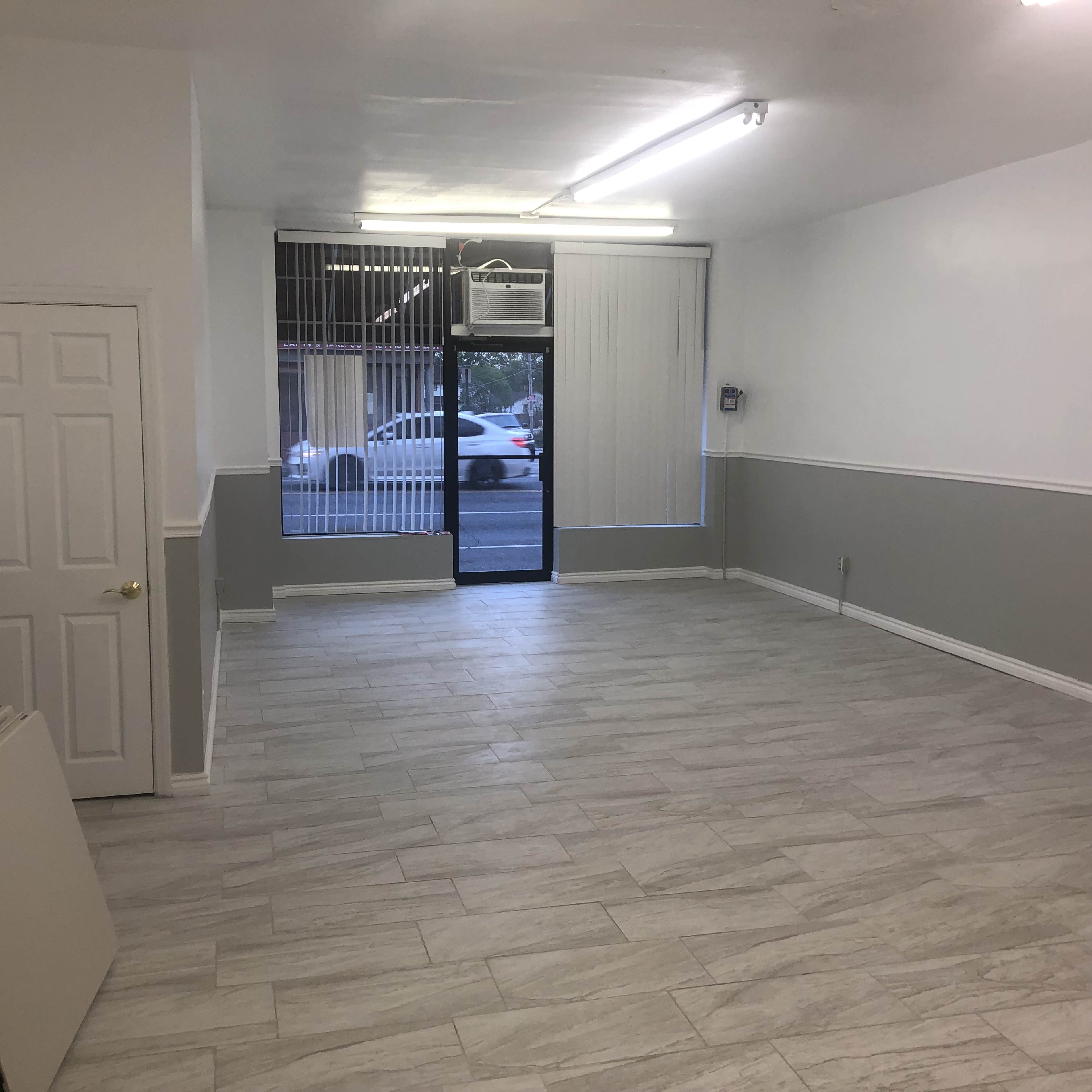 The image shows an empty interior space with gray and white walls, a tiled floor, and large windows facing a street.