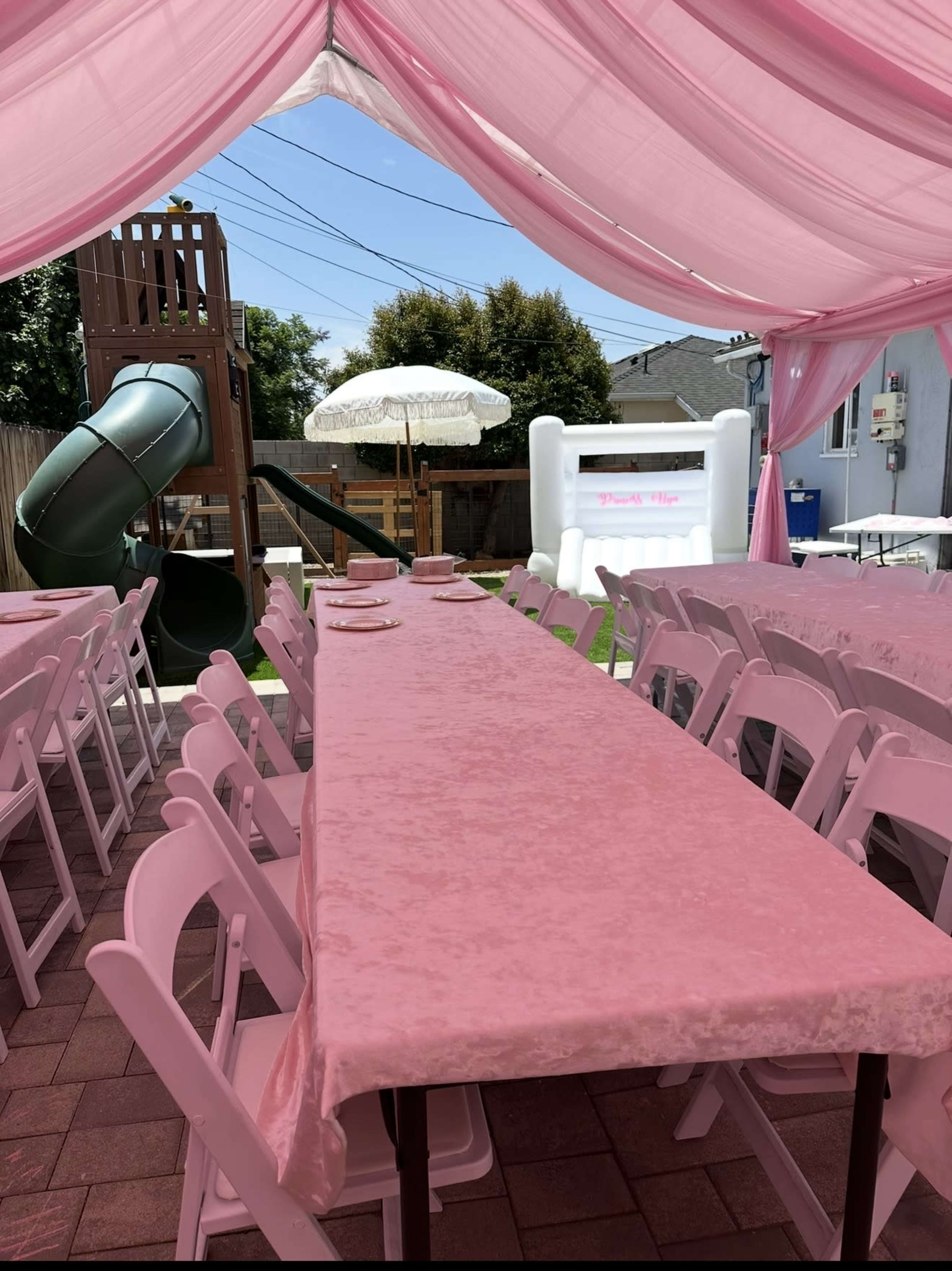 The image shows a setup for an outdoor event with a long pink table covered with a plush tablecloth, surrounded by rows of white chairs, under a tent with pink drapery, alongside a green slide and a white inflatable structure.
