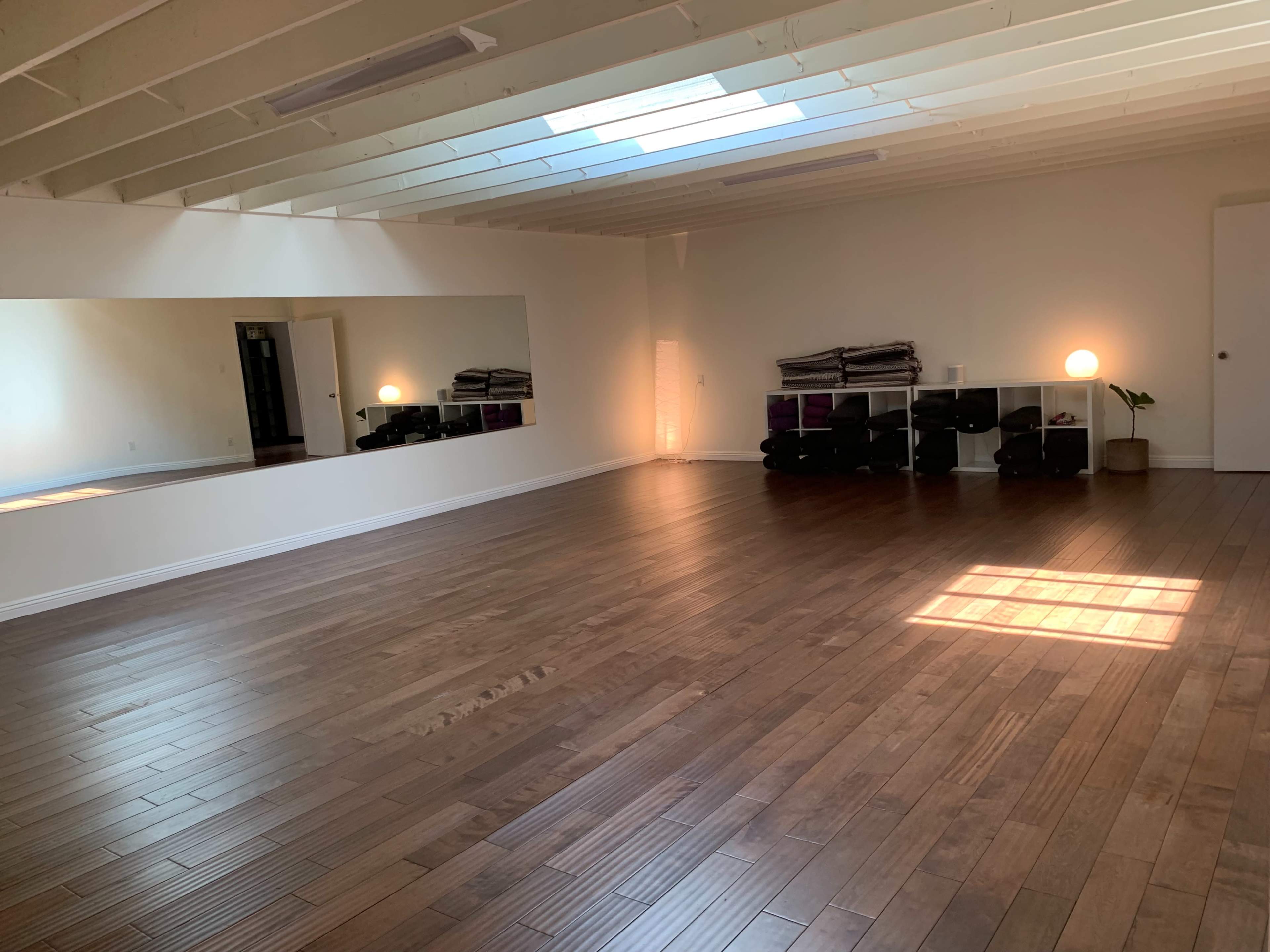 A spacious room with wooden floors, large mirrors on one wall, and neatly arranged shelves containing yoga mats and a lamp.