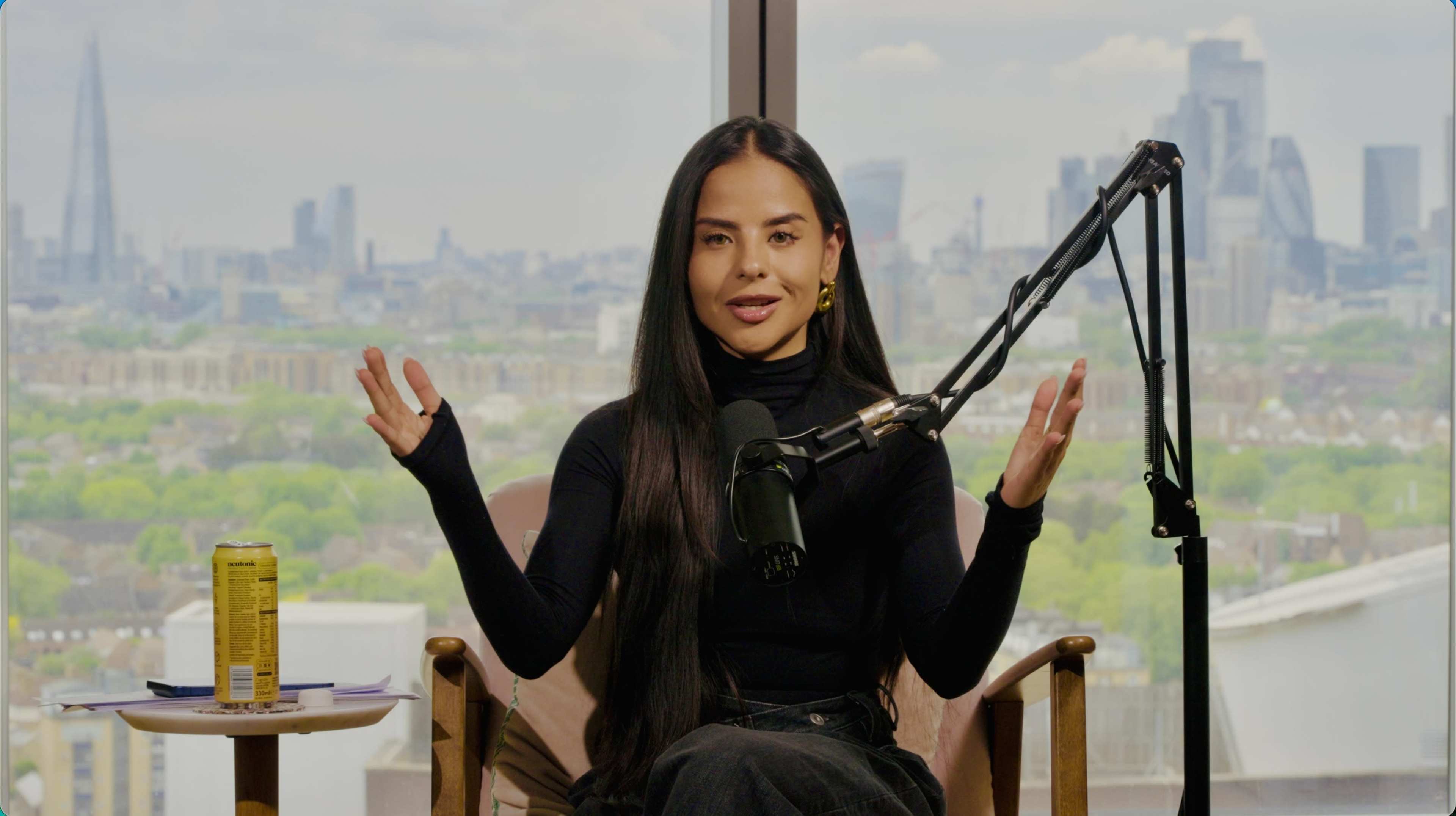 A woman with long black hair is seated in a chair with a microphone in front of her, gesturing while overlooking a city skyline through a large window.