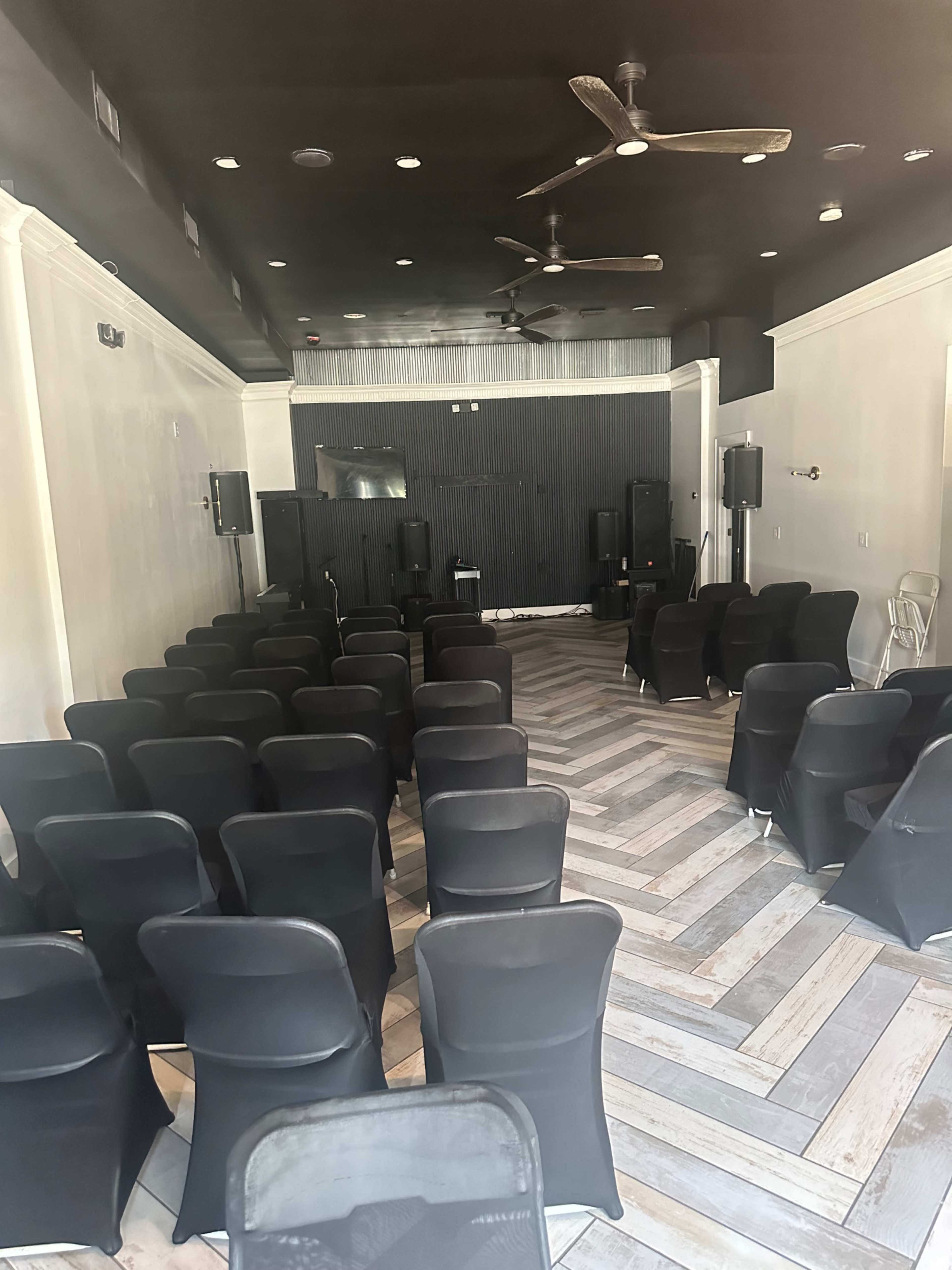 A room set up for an event with rows of black chairs facing a stage area, featuring audio equipment and a black backdrop.