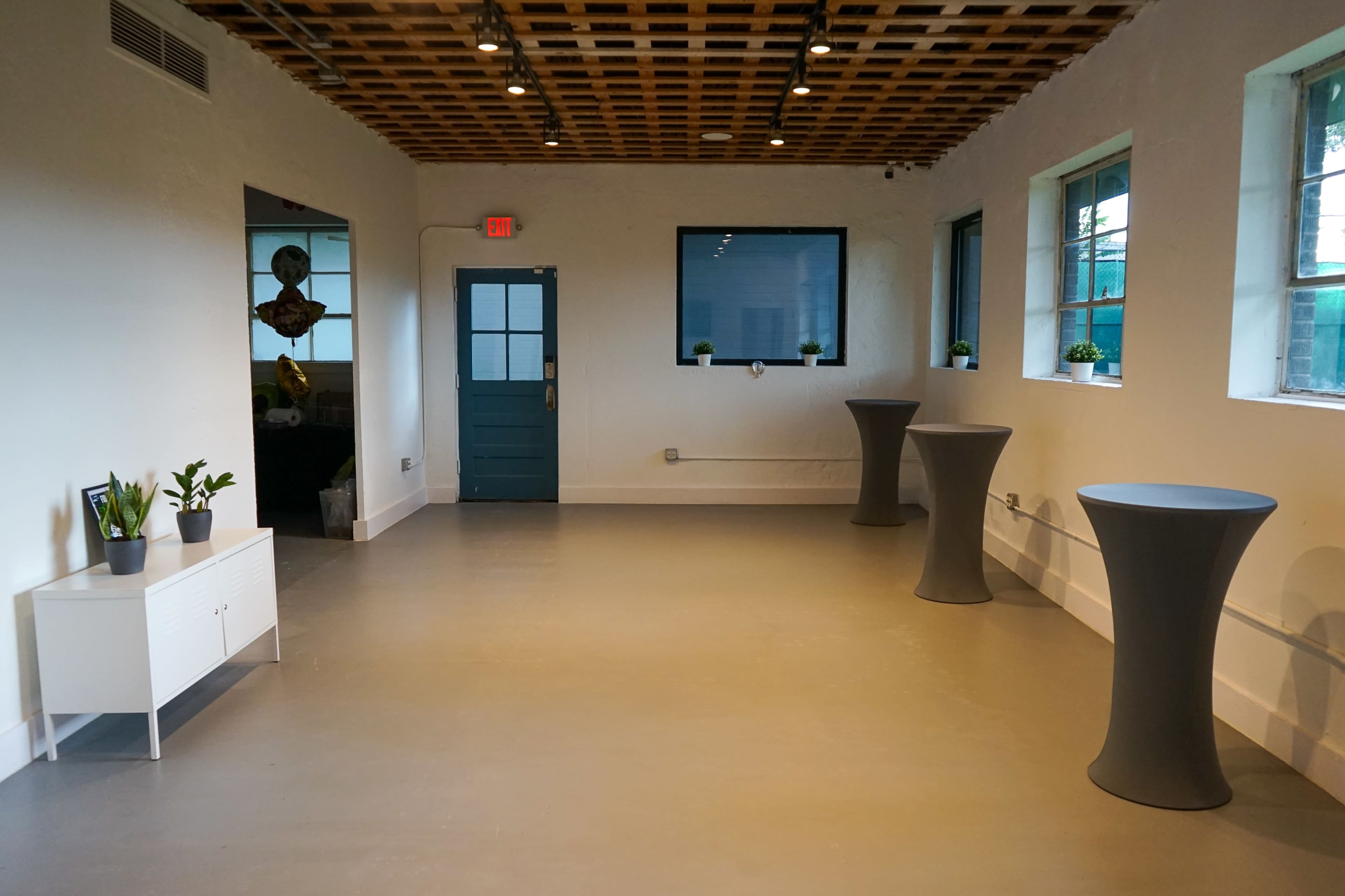 The image shows a modern, empty room with a wooden ceiling, large windows, and two tall tables arranged along the walls.