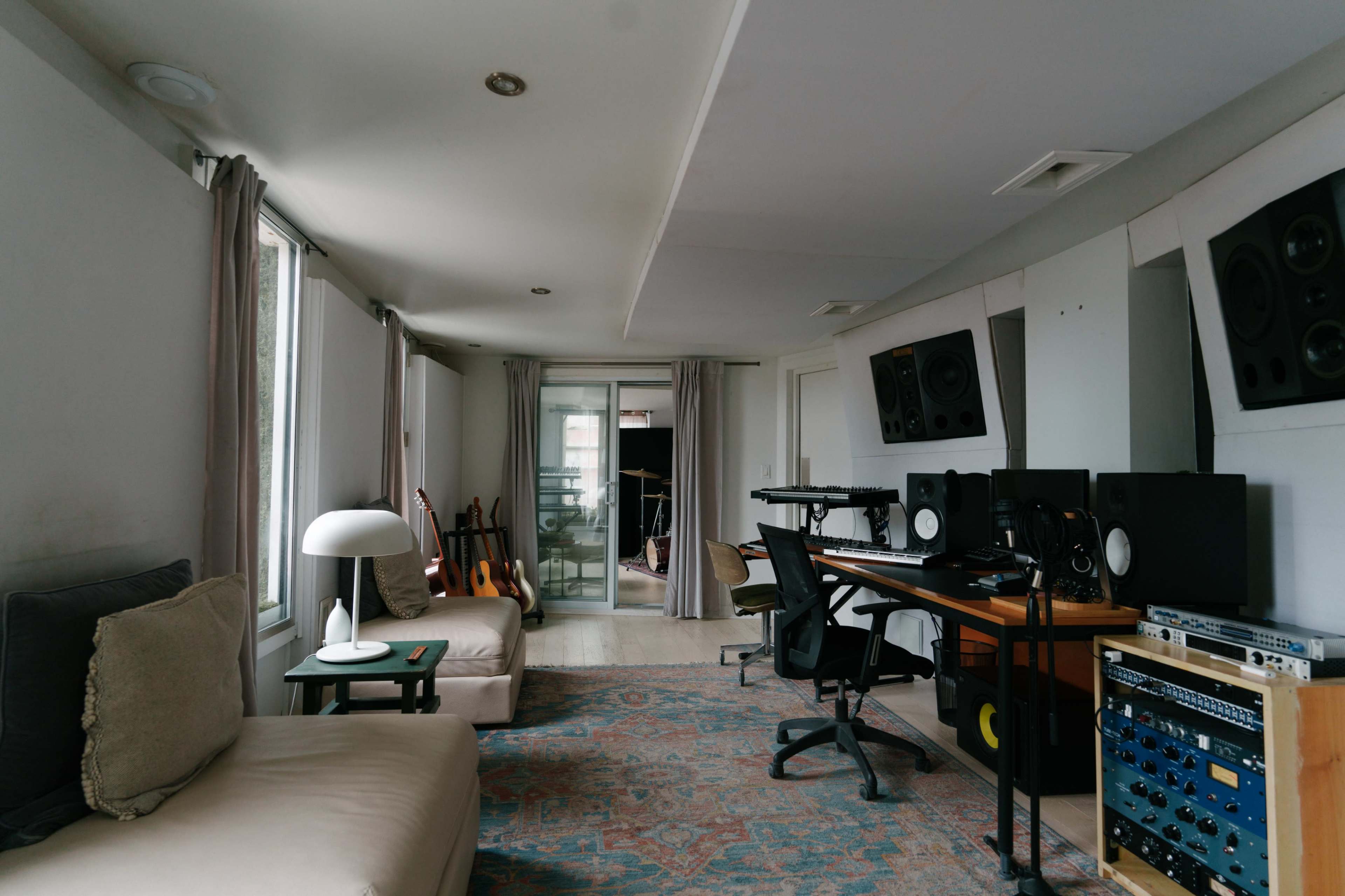 A music studio with a mixing desk, monitors, and instruments, featuring a spacious layout with large windows and modern furnishings.