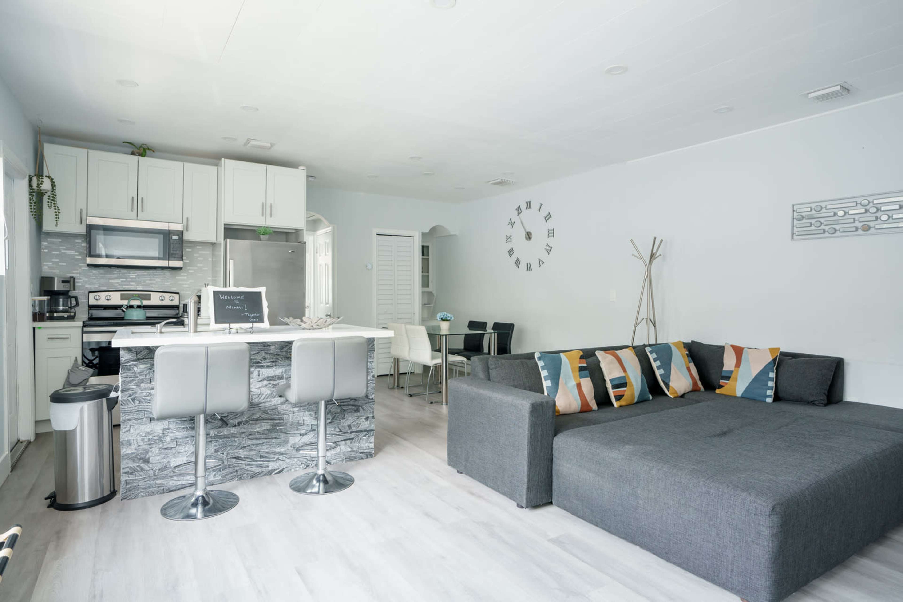 A modern, open-concept kitchen and living area with a gray sectional sofa, bar-style seating, and a dining table.