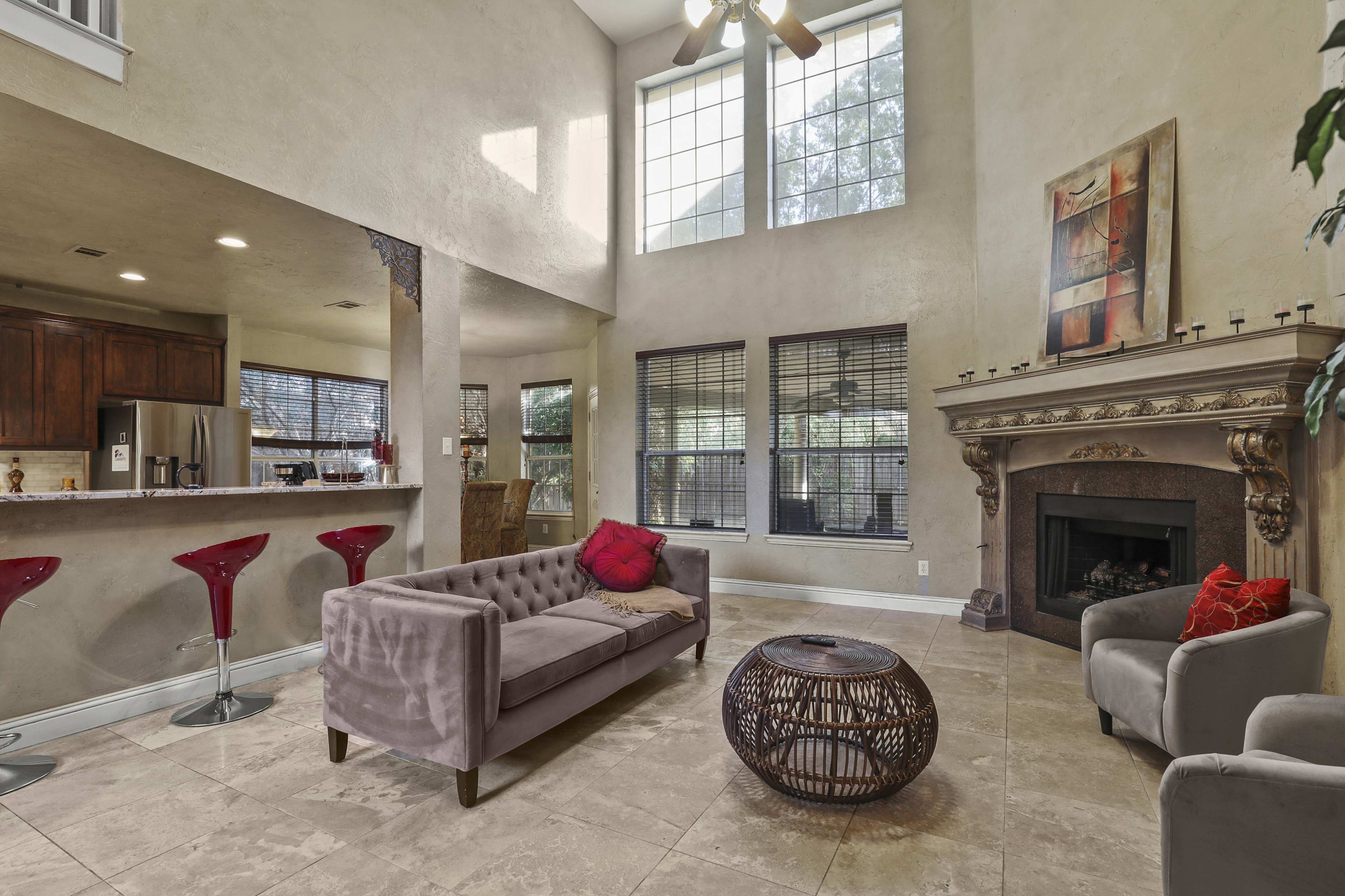 A spacious living area with a high ceiling, featuring a gray tufted sofa, a round wicker coffee table, and a fireplace surrounded by a decorative mantel, with a kitchen area visible in the background.