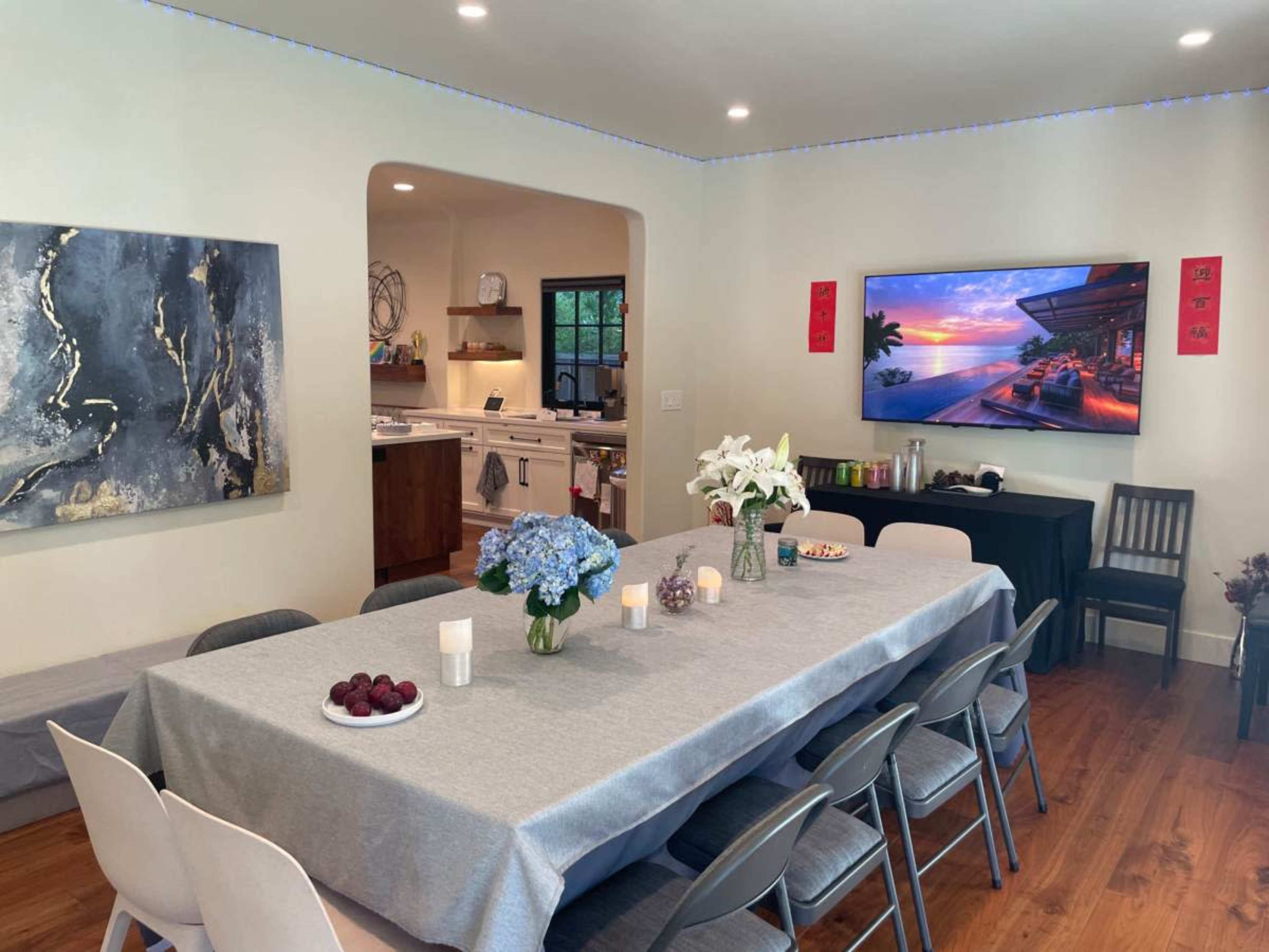 The image shows a dining area with a long table set for a meal, surrounded by chairs, with a modern kitchen visible in the background and a television mounted on the wall displaying a scenic image.