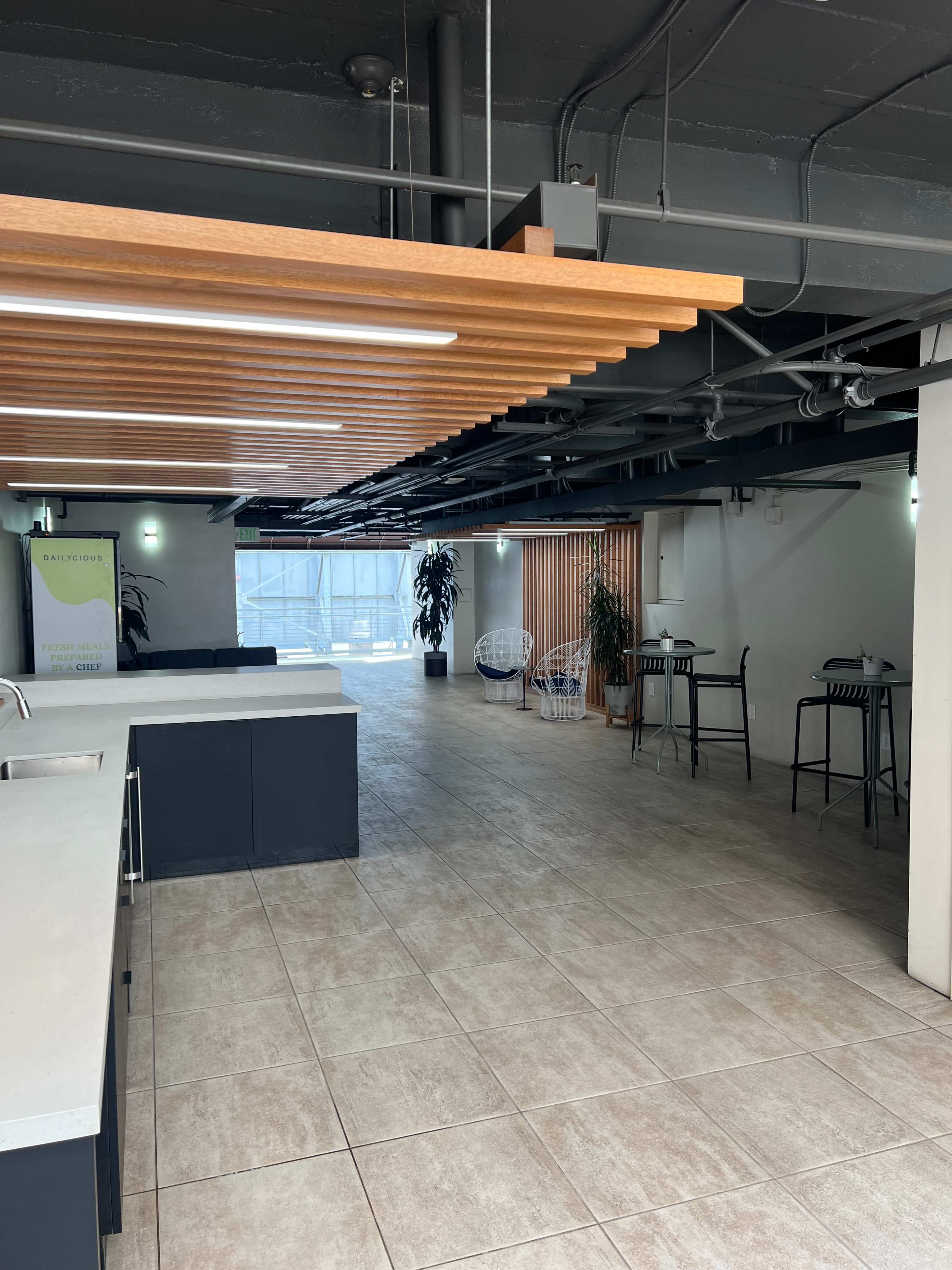 The image shows a spacious interior with a modern design, featuring a kitchen area on the left and tables in the background under a wooden ceiling.