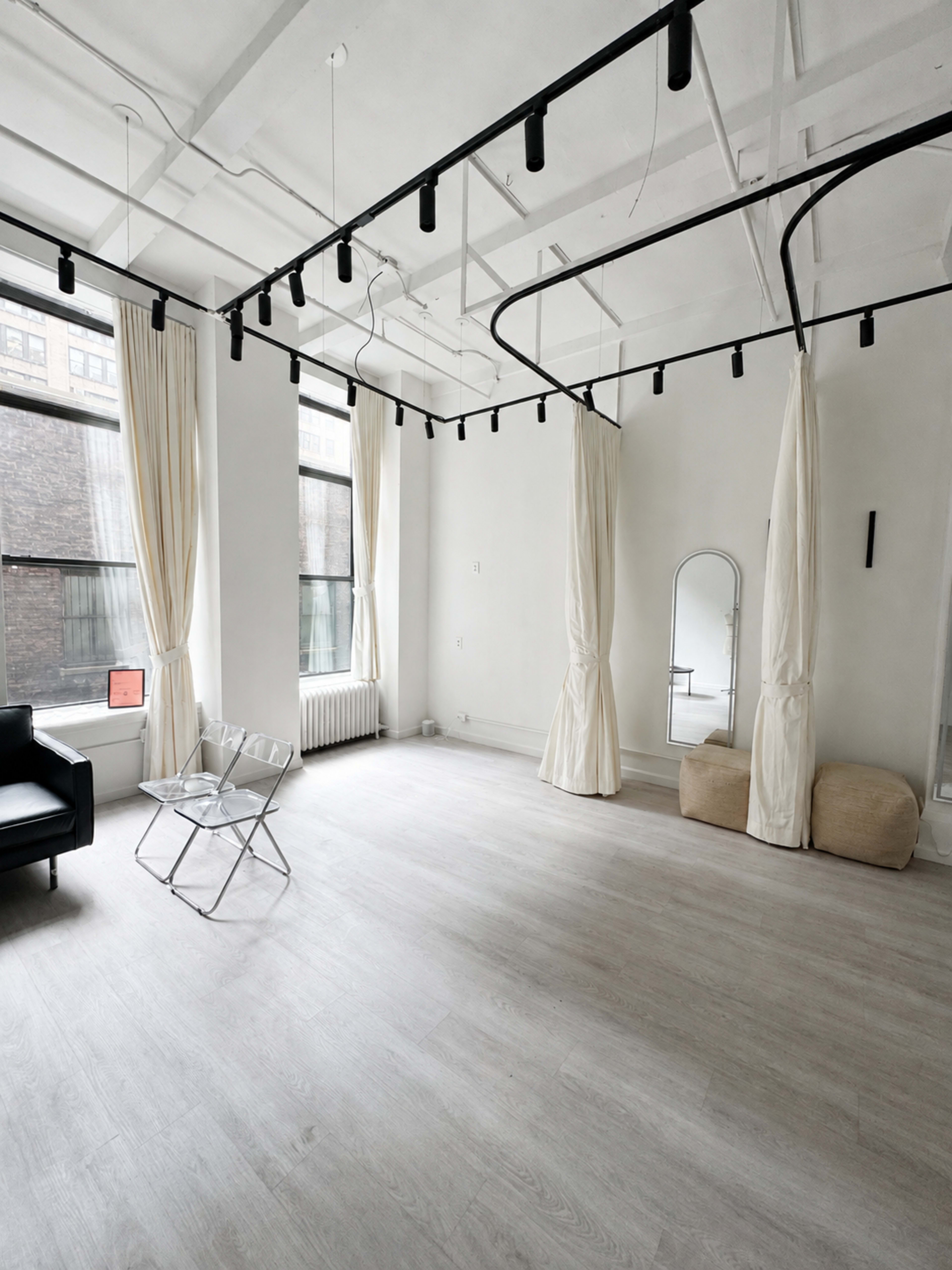 A bright, minimalistic room features large windows, white walls, and light wood flooring, with a black chair, a metal chair, and two fabric poufs.
