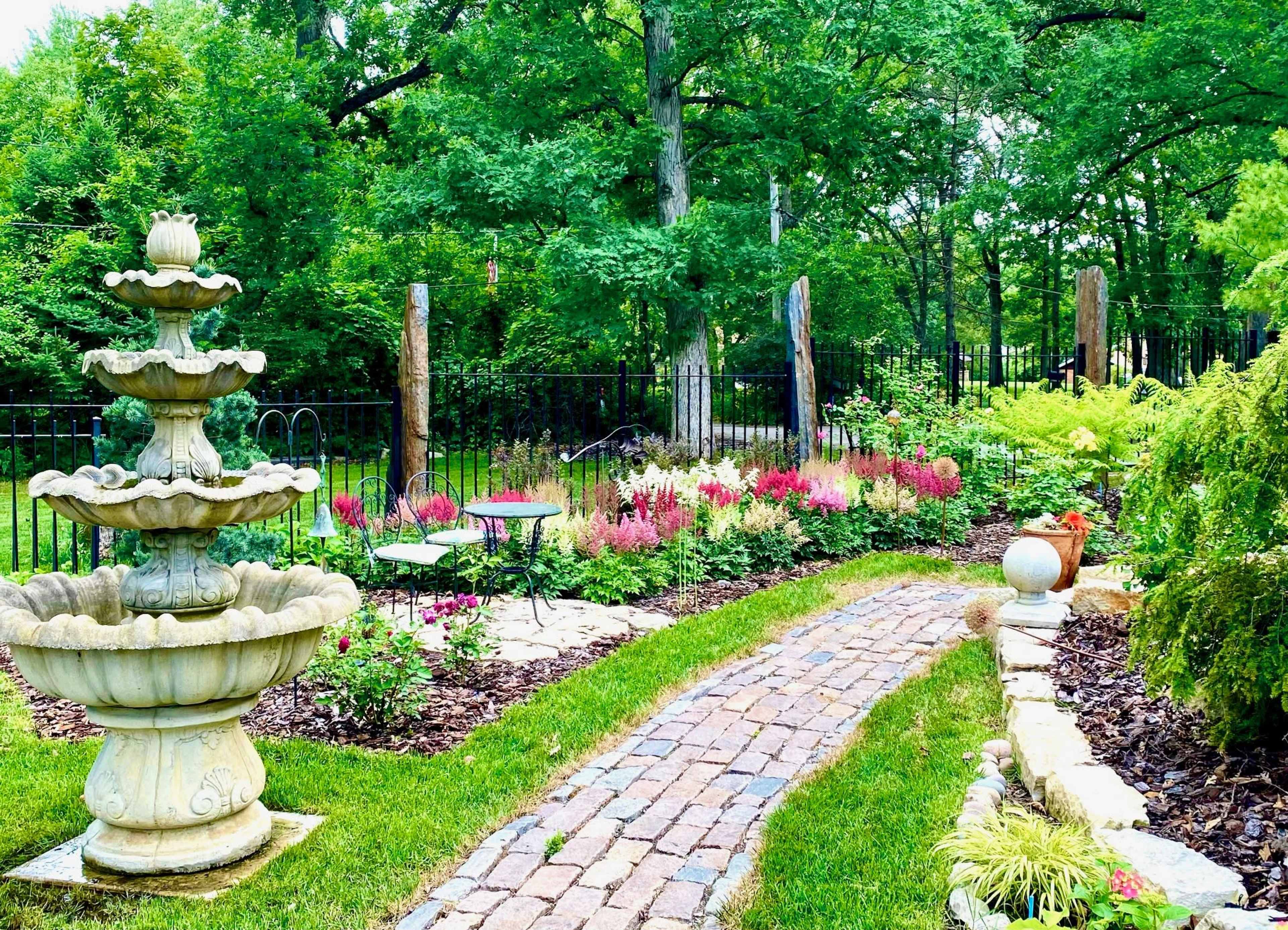 A stone pathway winds through a garden featuring a fountain and colorful flower beds.