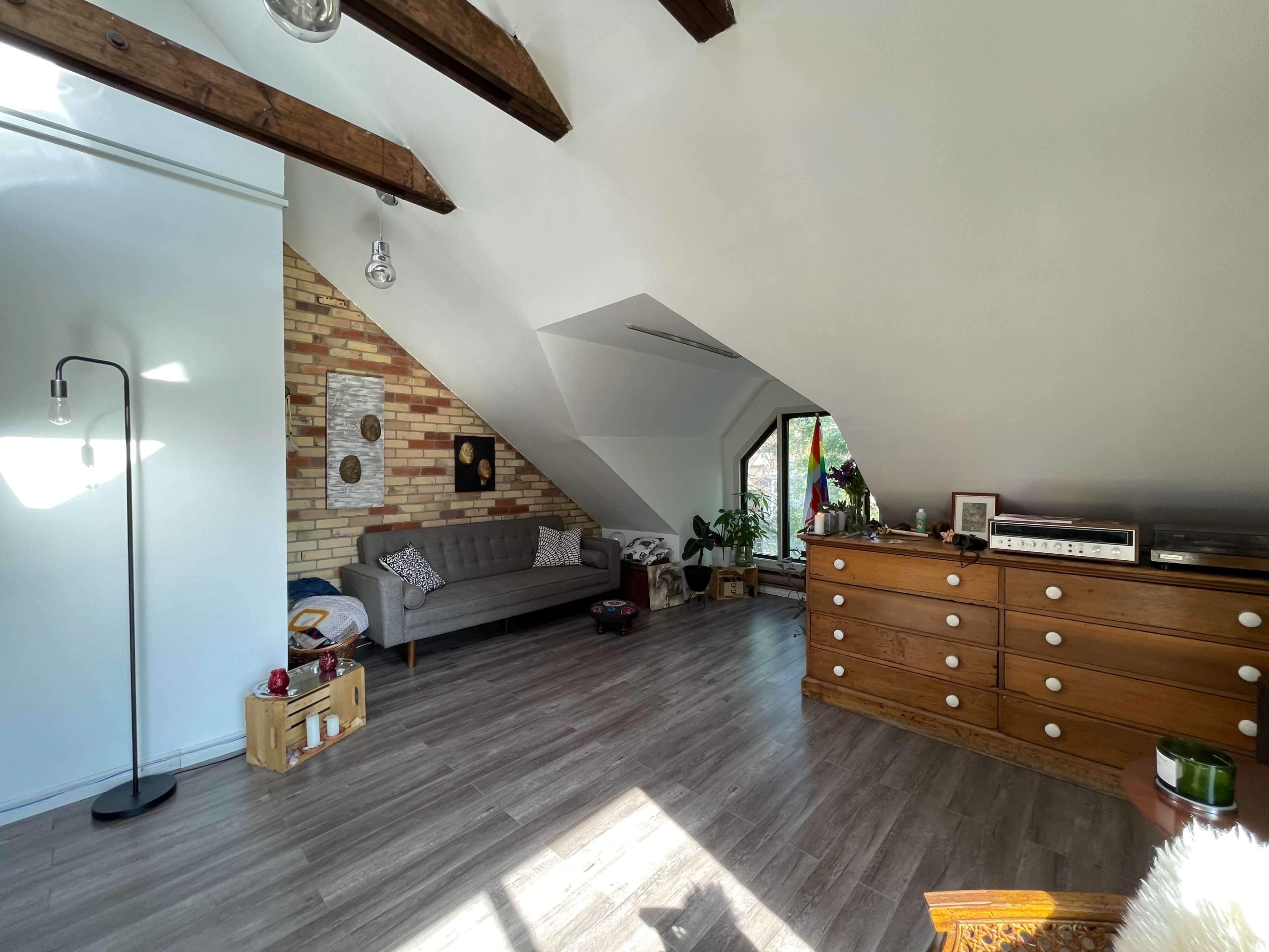 An attic room featuring a gray couch, a wooden dresser, and large windows with natural light illuminating the space.
