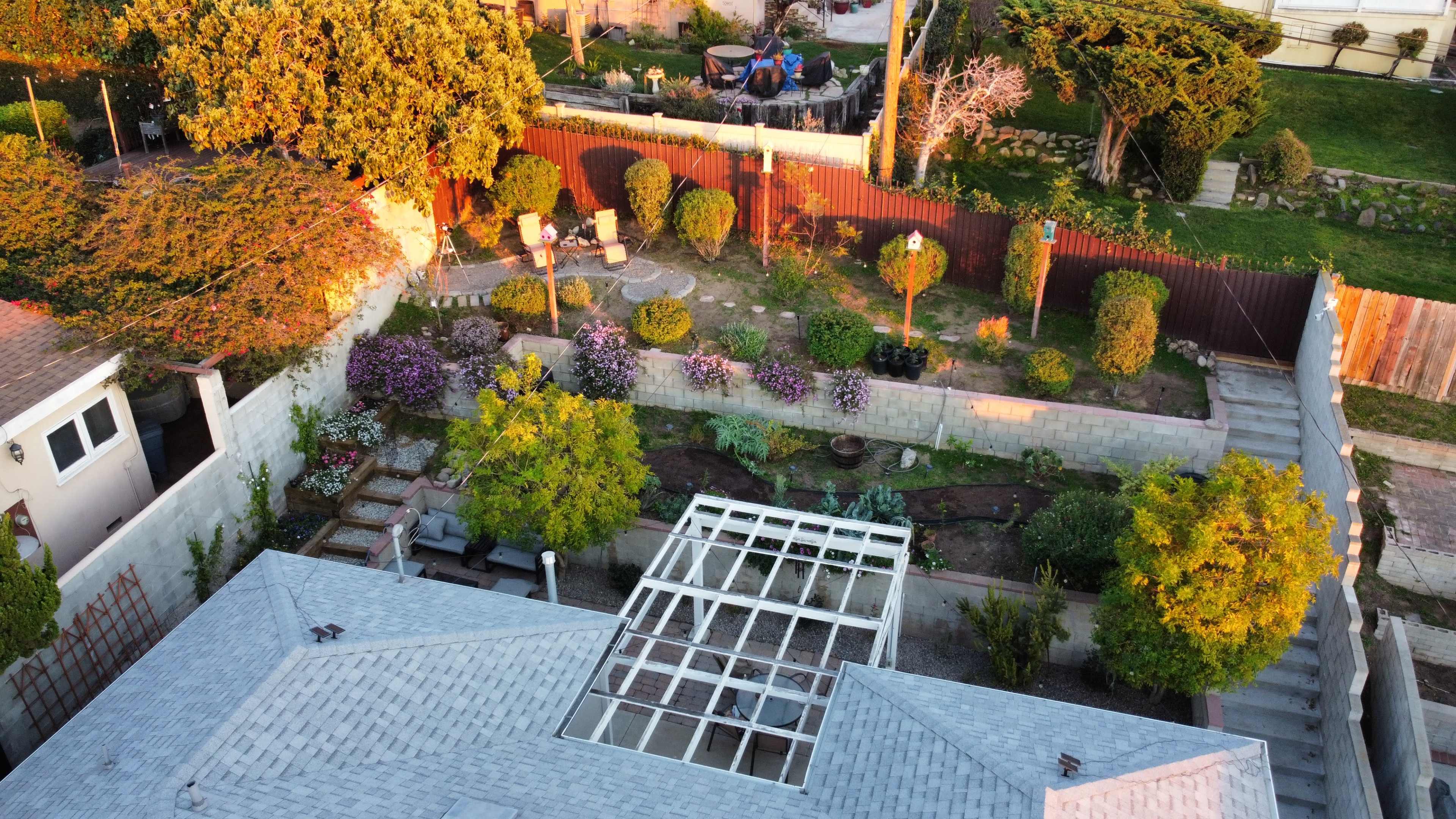 The image shows an aerial view of a garden with well-maintained shrubs, flower beds, and seating areas surrounded by a fence.
