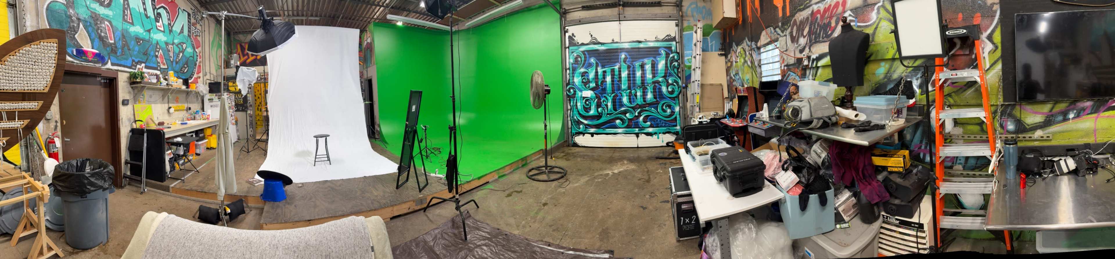The image shows a panoramic view of a creative studio featuring a green screen area, a white backdrop, a stool, lighting equipment, and various art supplies and clutter along the walls.