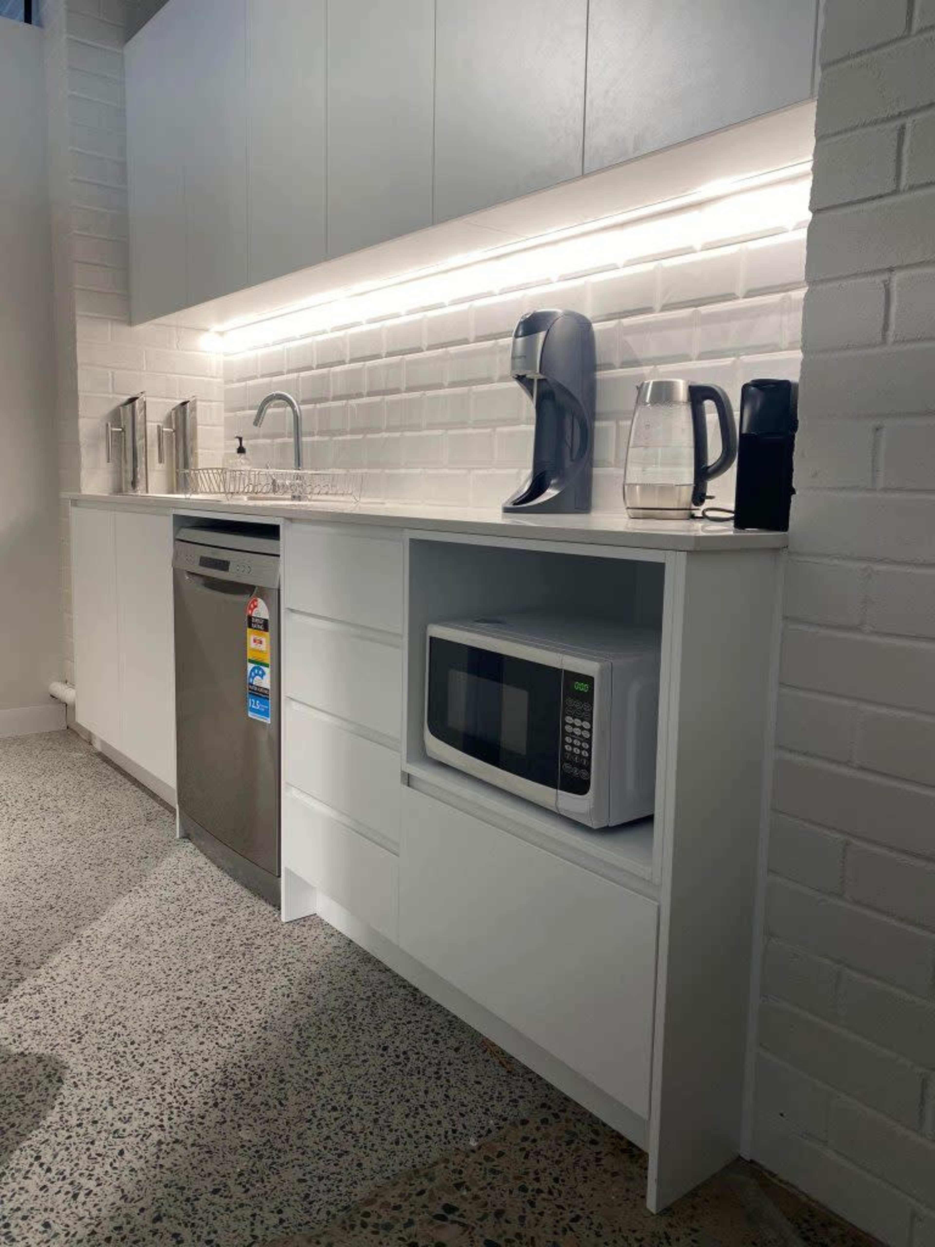 A modern kitchen area with white cabinetry, a stainless steel dishwasher, a microwave stored in a cabinet, and countertop appliances including a coffee maker and a kettle.