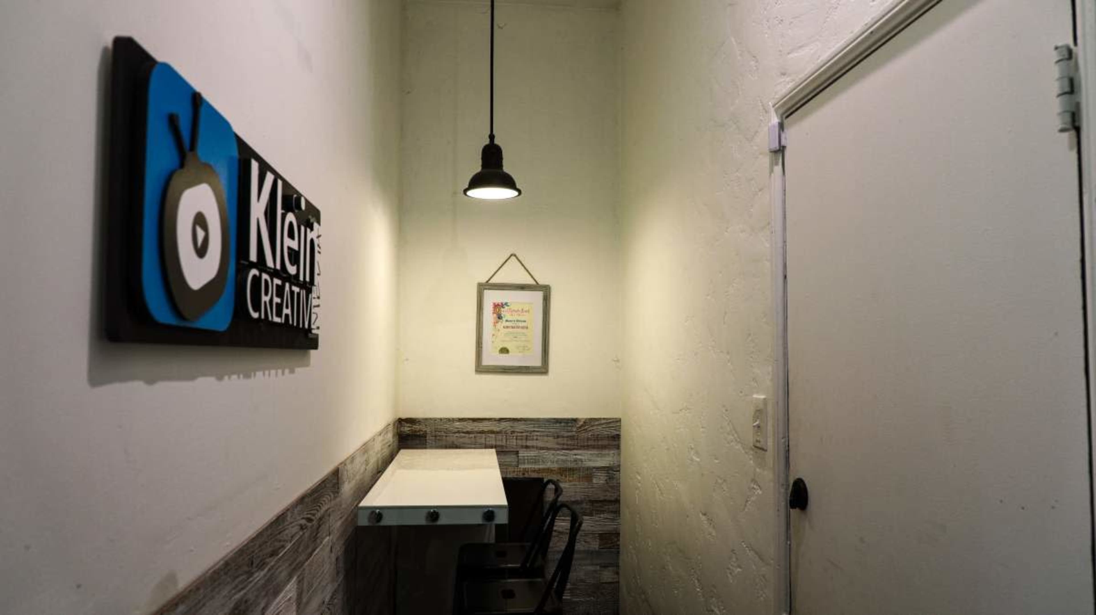 The image shows a narrow hallway with a sign for "Kleir Creative" on the wall, a framed document hanging, and a small table with a chair.