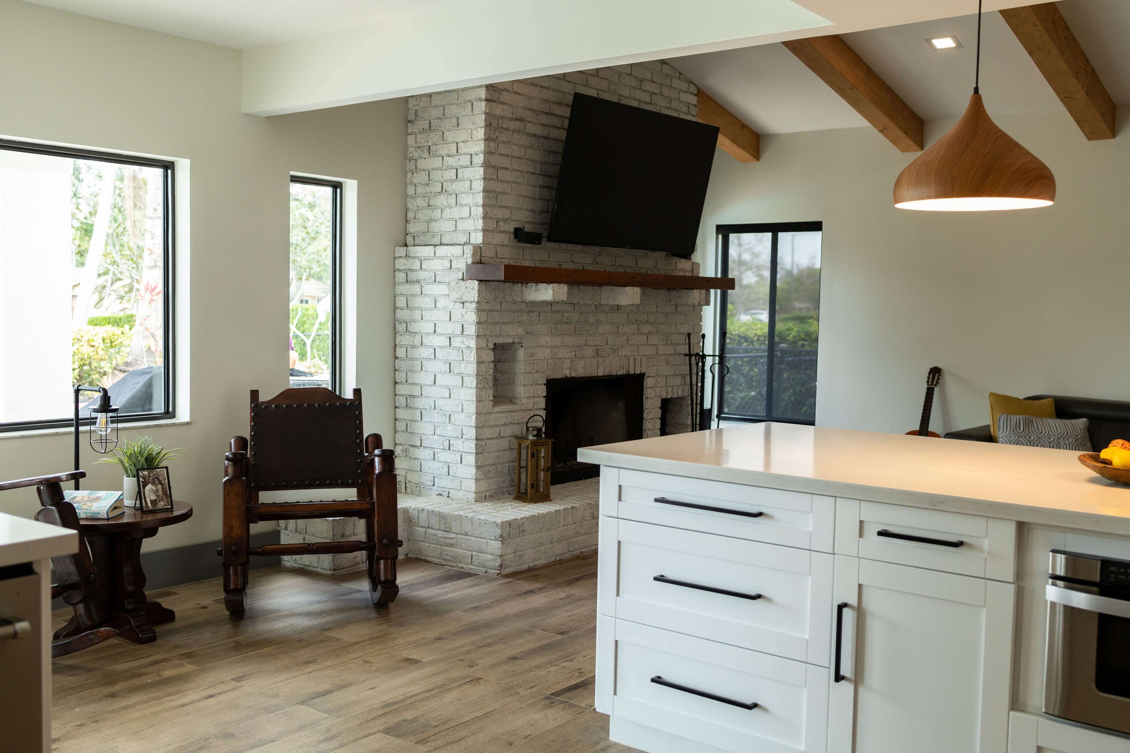 A modern living room with a brick fireplace, a television mounted above it, and an open kitchen area featuring white cabinets and a wooden dining table.